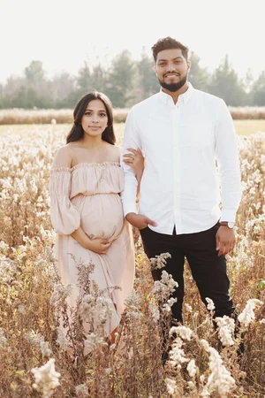 An expectant couple stand arm in arm in a field of tall dried wildflowers, her hands on her bump, both smiling at the camera.