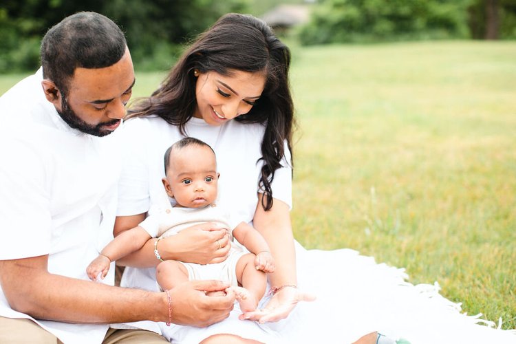 A mother and father sit close on a blanket in a meadow, smiling down at their baby son on her lap.