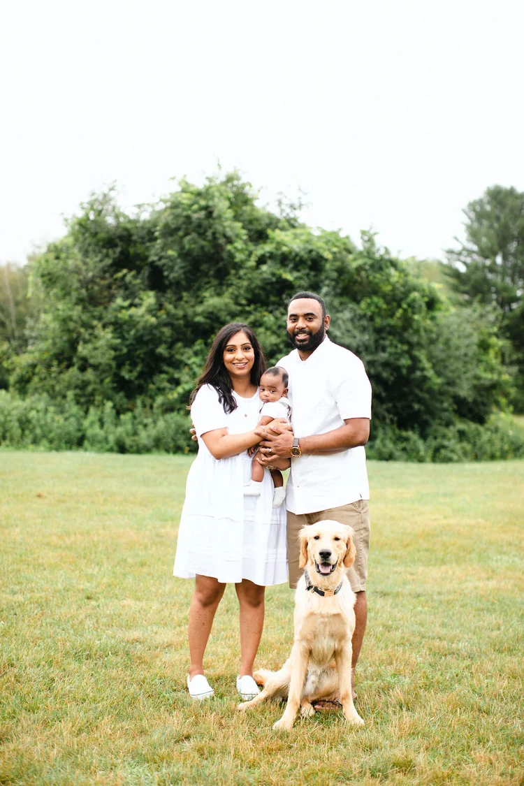 A mother and father in white stand in a meadow holding their baby, their golden retriever sitting at their feet.