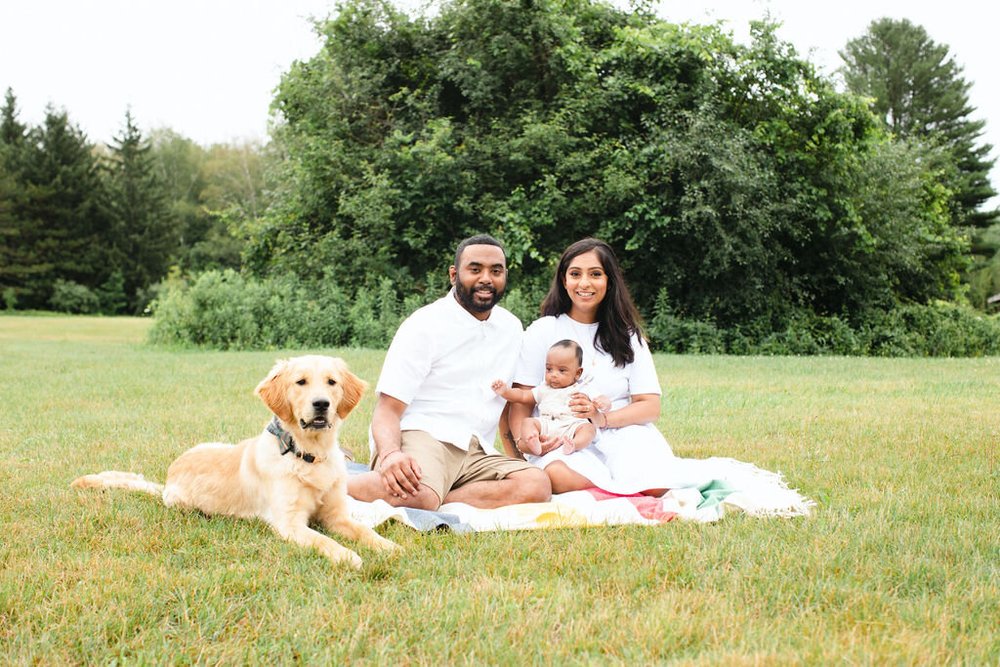 A mother, father, baby and golden retriever sit together on a striped blanket in a wide green meadow.