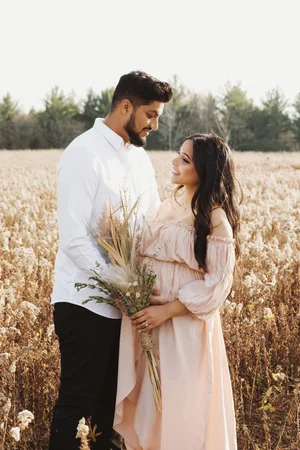 An expectant couple smile at each other in a field of dried wildflowers as she holds a bouquet of dried grasses against her bump.
