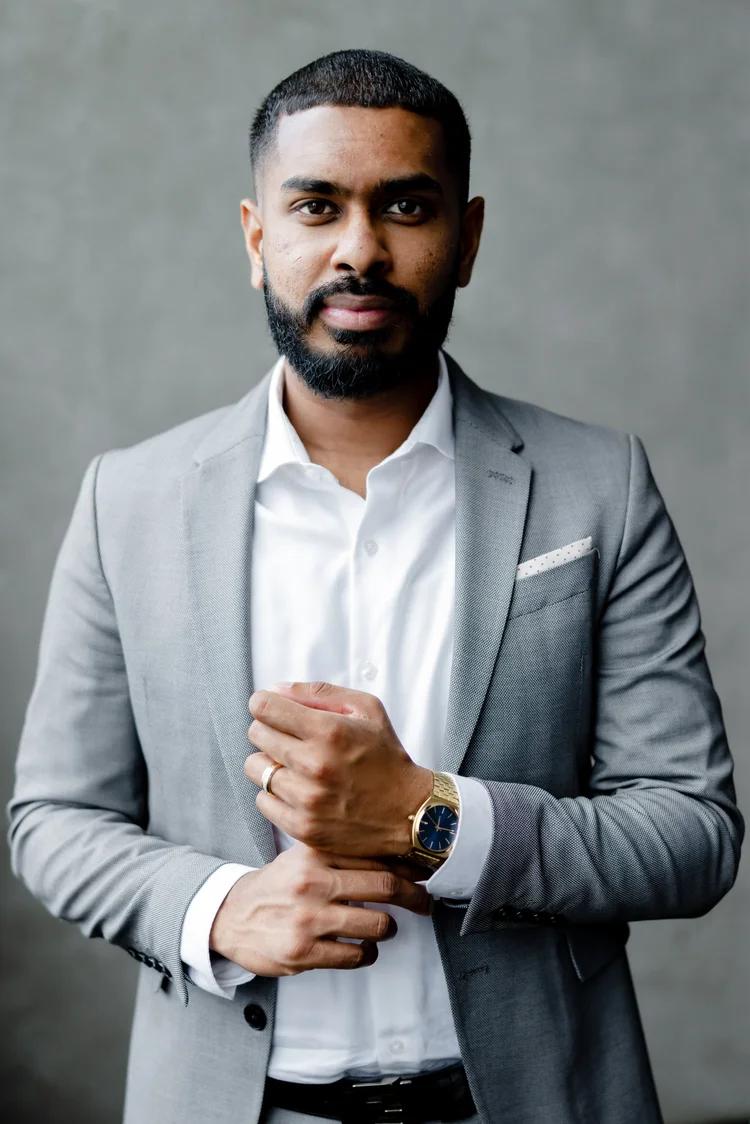 A bearded man in a grey suit adjusts his cuff against a plain grey studio backdrop.