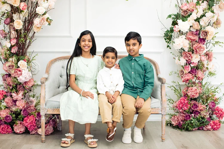 Three siblings, a girl and two boys, sit smiling on a grey settee between columns of pink and cream roses.