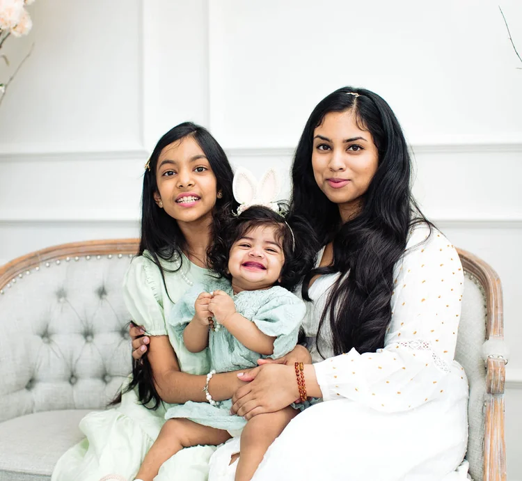 A mother sits on a grey tufted settee with her two daughters, the baby laughing on her big sister’s lap.
