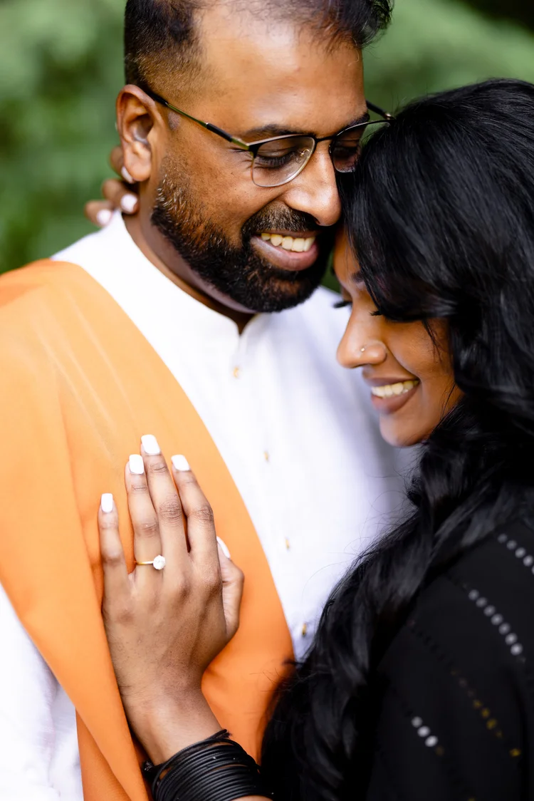 A bearded man in a white kurta and orange shawl smiles as a woman in black rests her face against his, her ringed hand on his chest.