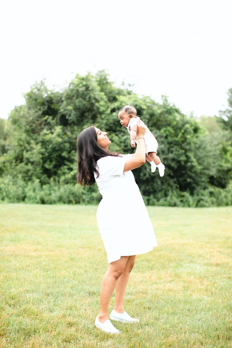 A mother in a white dress lifts her baby son high above her head in a green meadow, smiling up at him.
