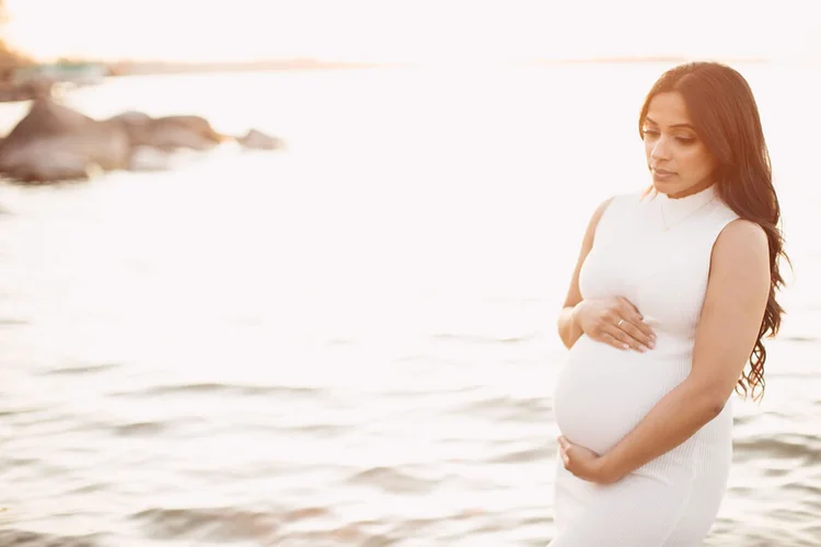 A pregnant woman in a fitted white dress stands in shallow lake water, eyes lowered, her hands on her bump.