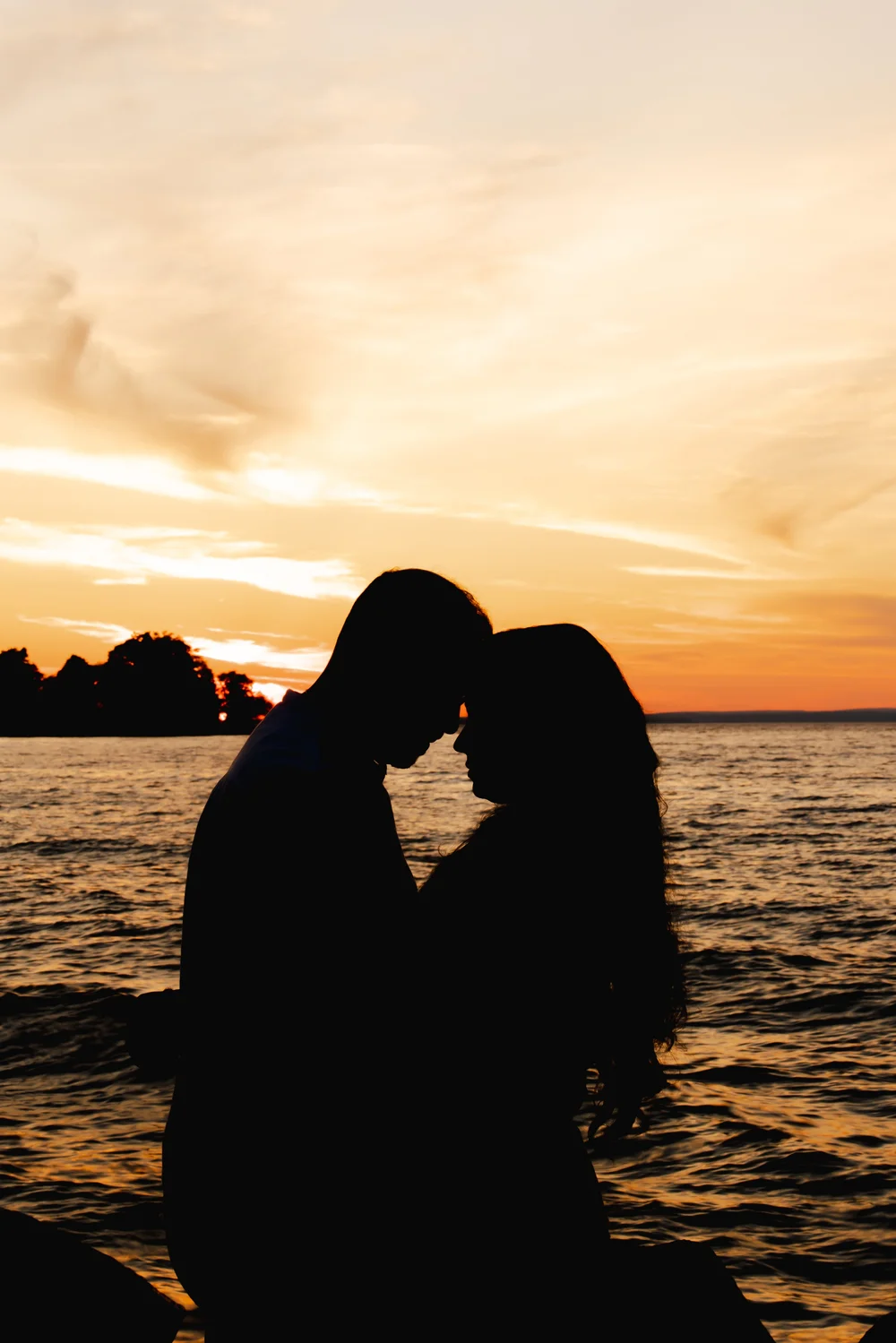 A couple in silhouette stand forehead to forehead against an orange sky over open water.