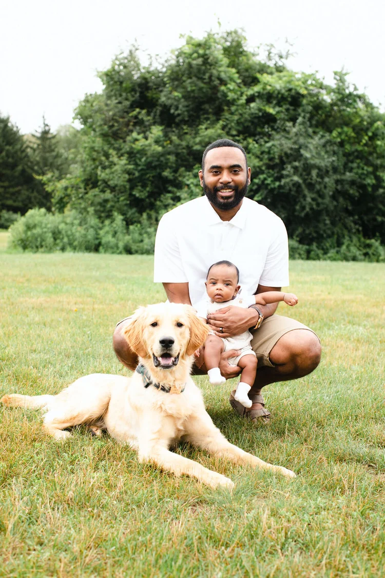 A smiling father crouches in a meadow with his baby son on his knee, their golden retriever lying in the grass in front.