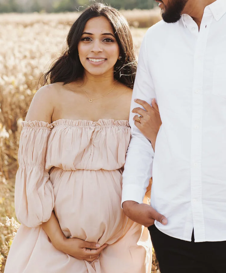A smiling pregnant woman in a blush off-shoulder dress holds her bump with one hand and her partner's arm with the other.