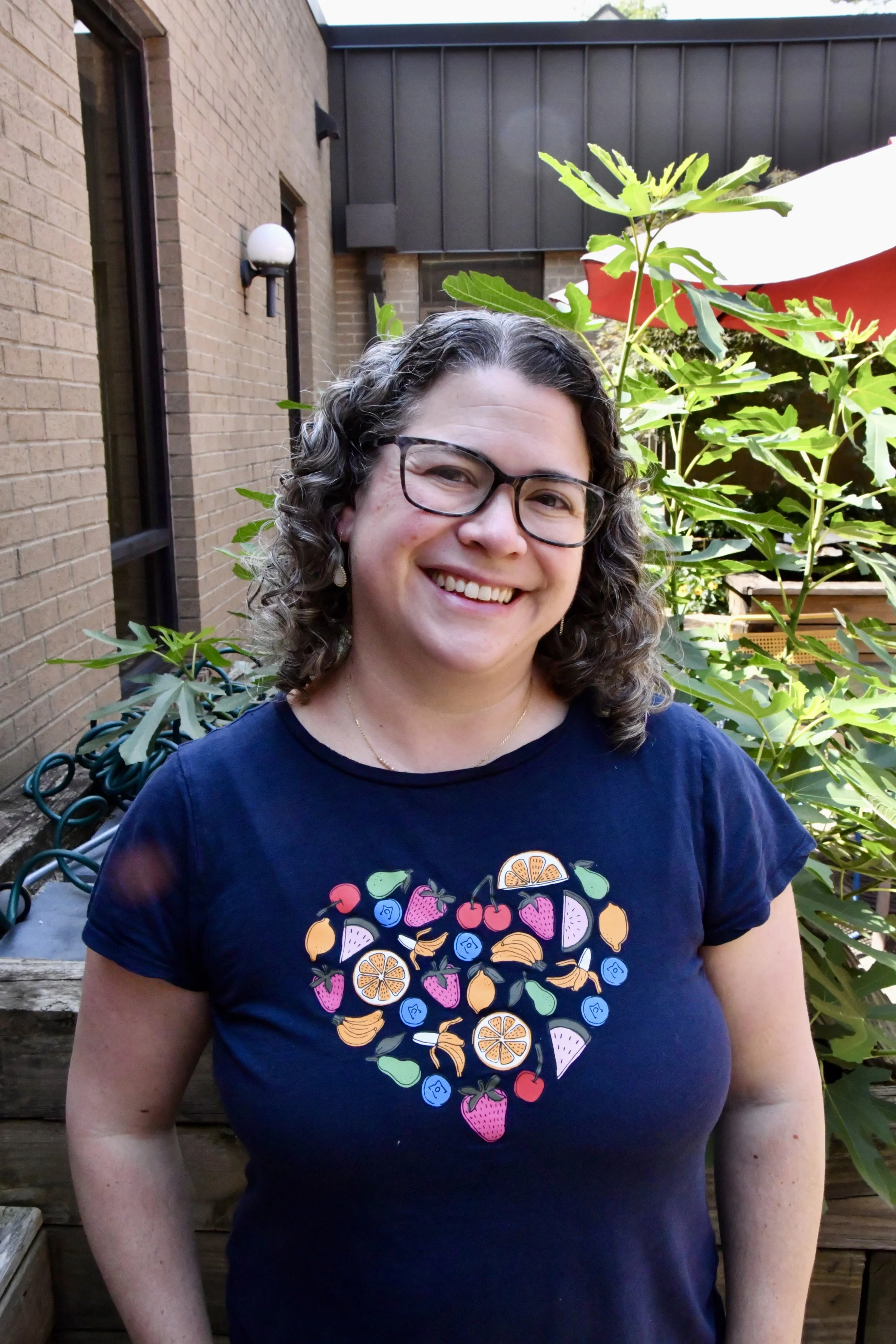 Close-up of a smiling woman with glasses, dark hair, wearing a floral sleeveless top, outdoors with green foliage in the background.