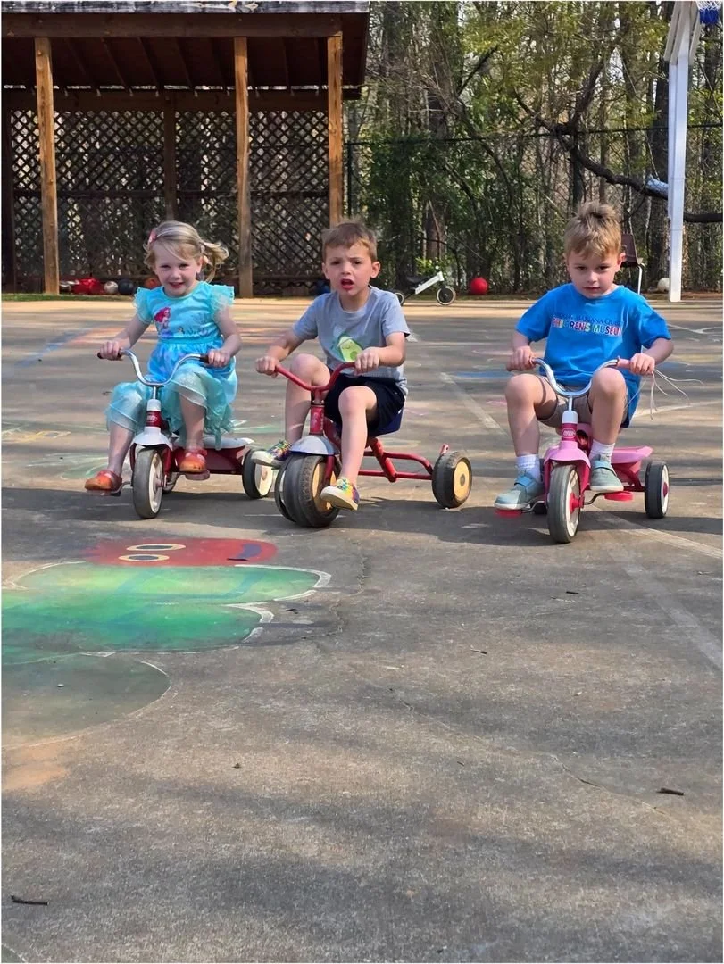 Three young children riding tricycles on a paved backyard area with chalk drawings, a wooden deck or shed in the background, and trees beyond.