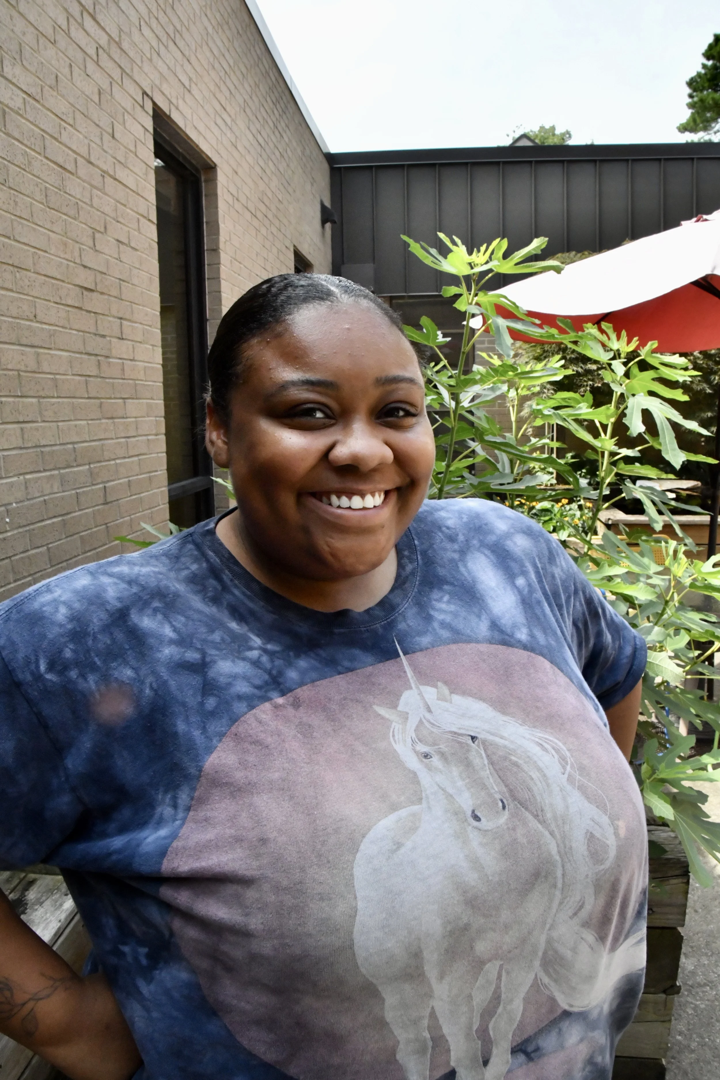 Close-up of a smiling woman with short curly black hair, wearing a gray shirt, outdoors in a garden setting.