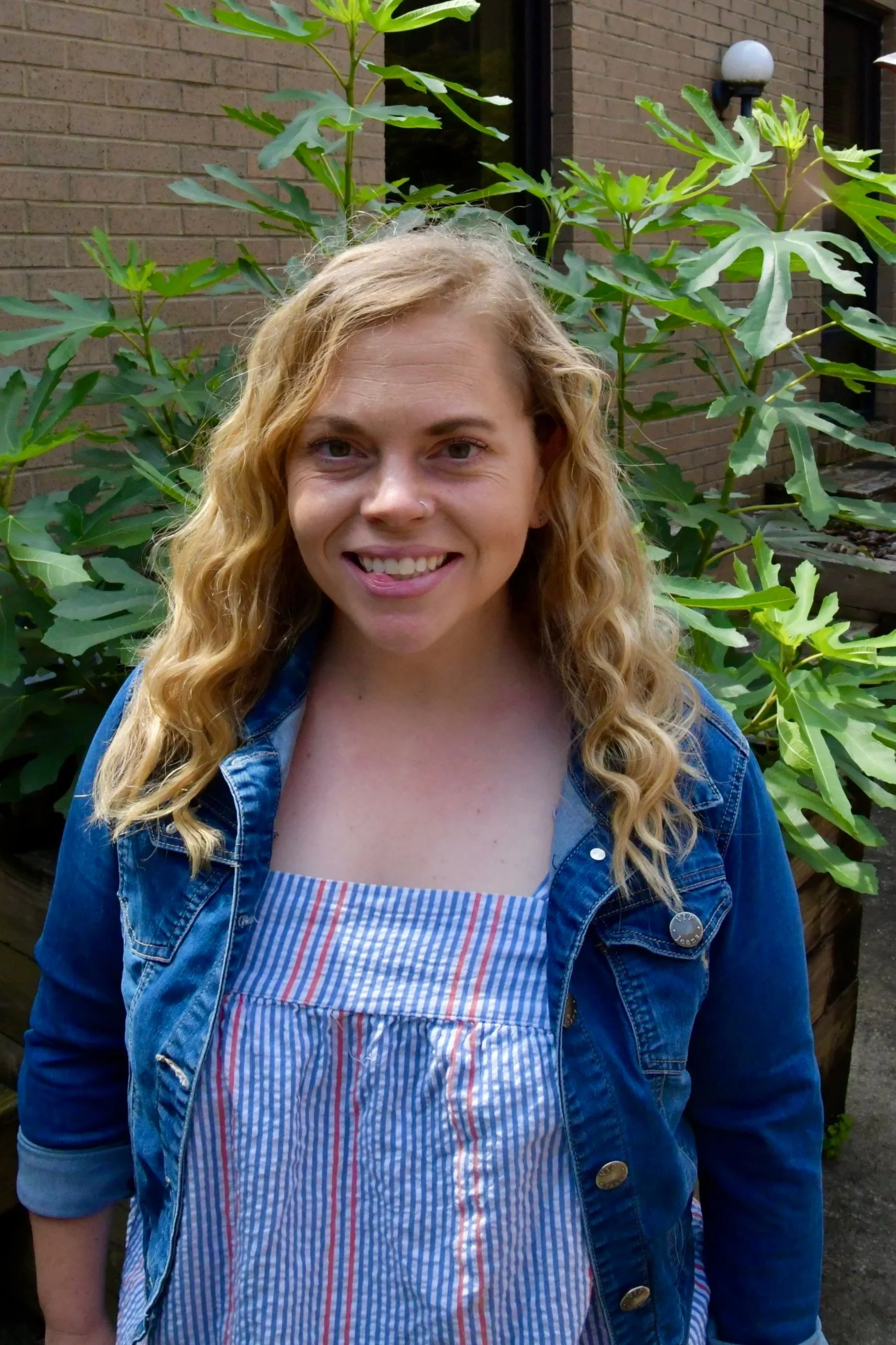 A woman with long blonde hair smiling outdoors in front of greenery, wearing a blue top.