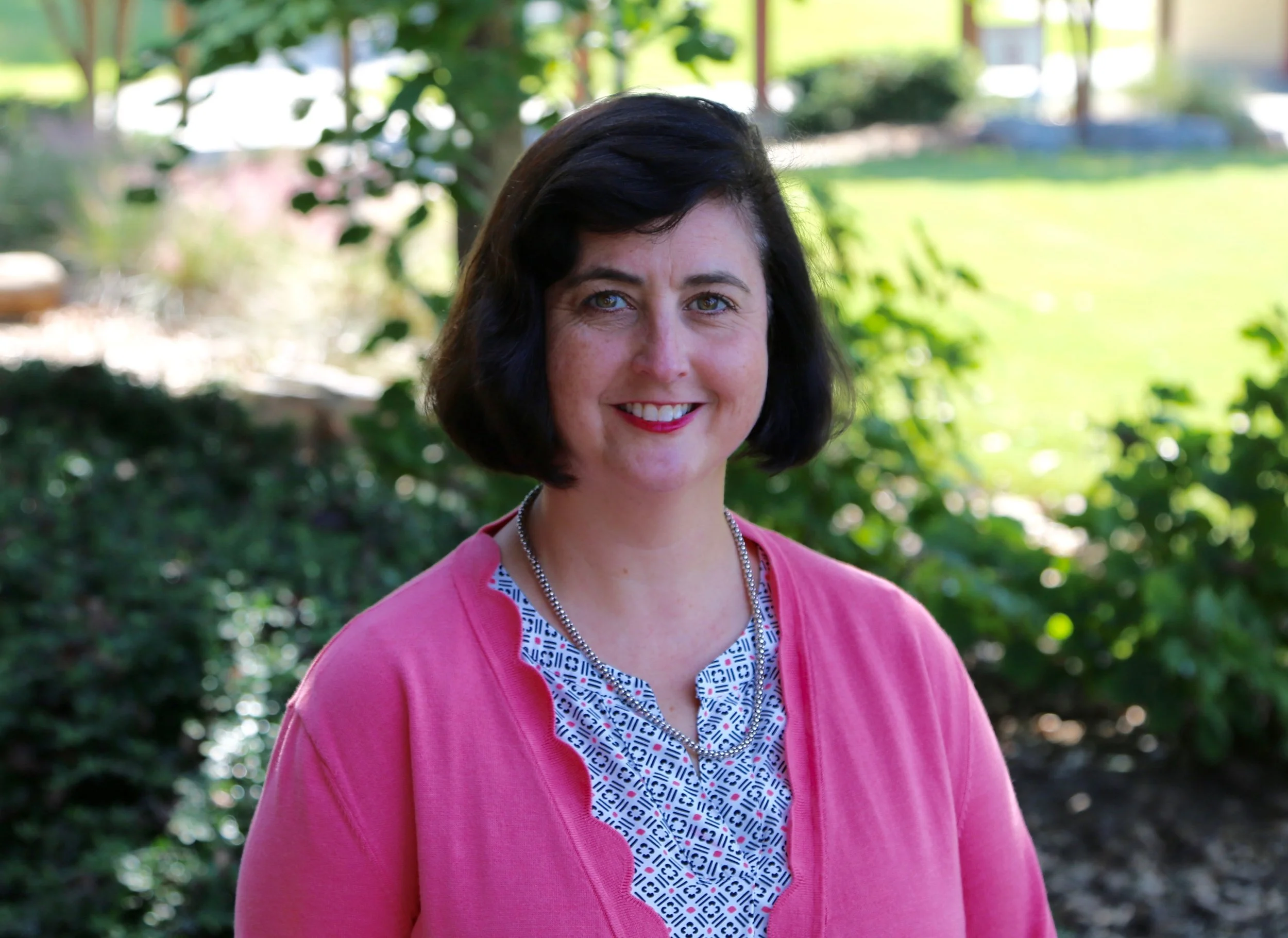 A woman with long blonde hair, blue eyes, and a bright smile, standing outdoors in front of green foliage, wearing a black sleeveless top, a necklace with a heart-shaped pendant, and a fitness tracker on her wrist.