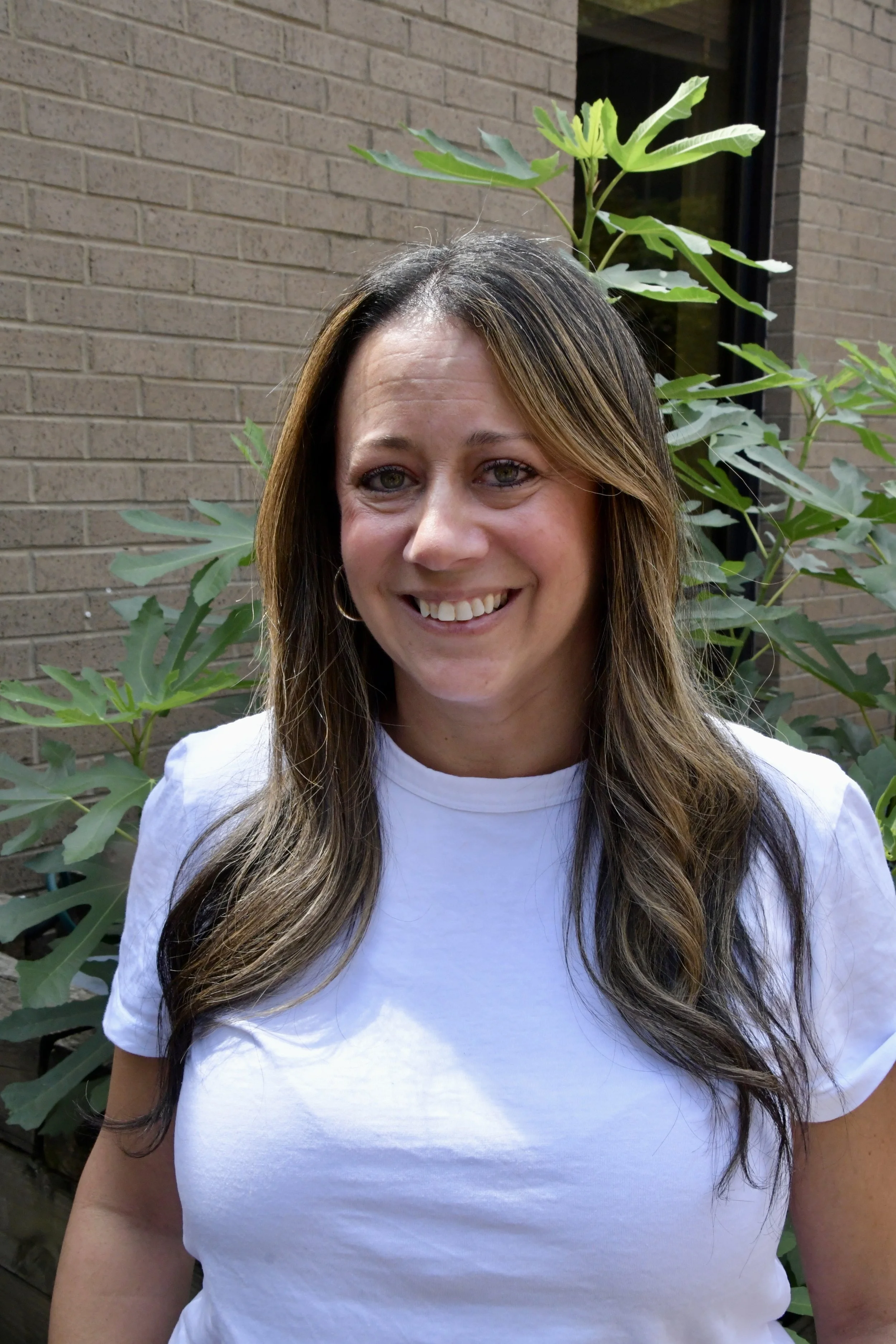 Smiling woman with long brown hair and green eyes outdoors with green foliage in the background.