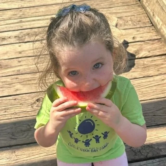 A young girl with curly hair, wearing a bright green shirt, is eating a slice of watermelon outside on a wooden deck.
