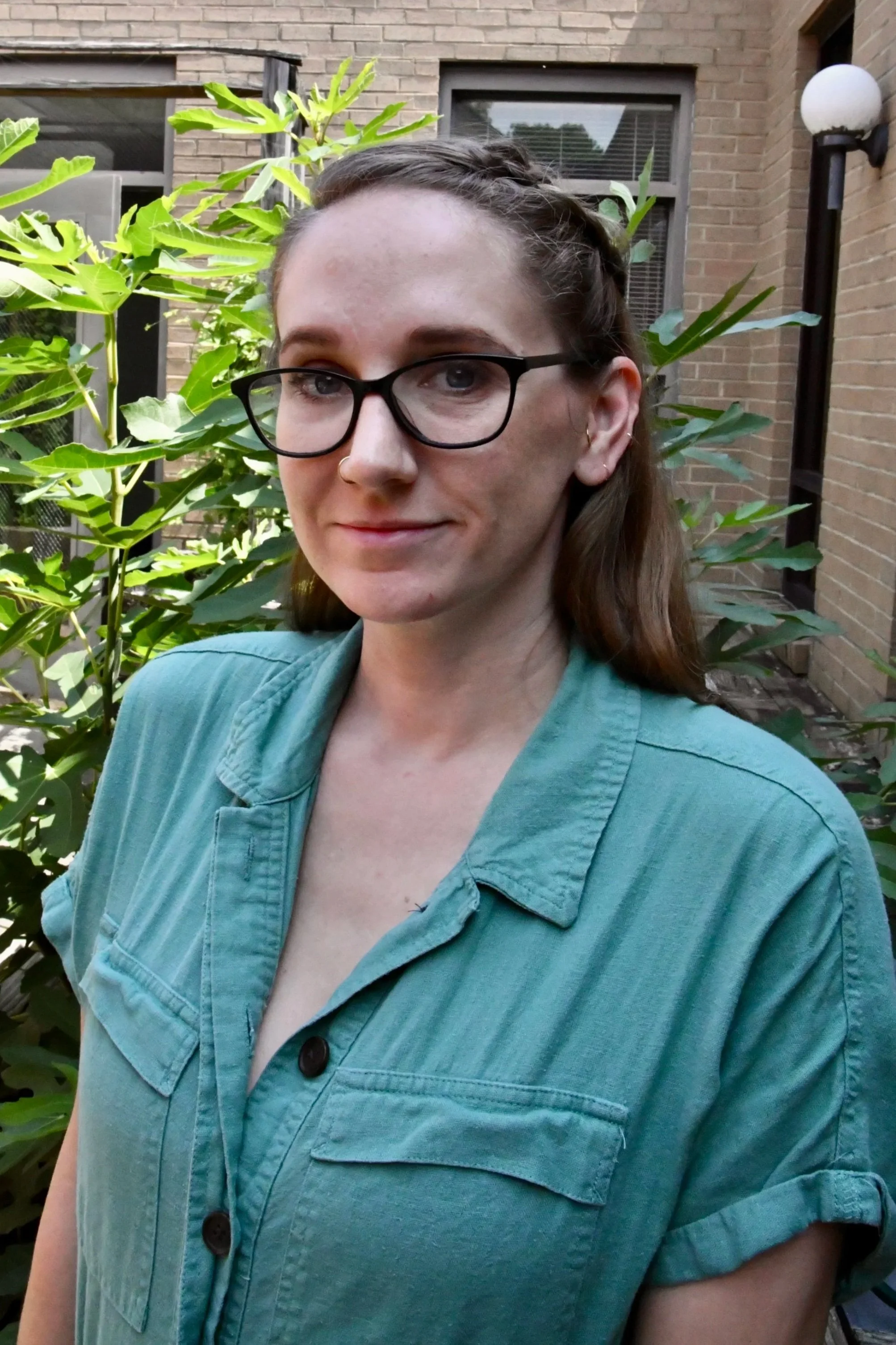 A young woman with glasses and a nose ring outdoors, with blurred green and yellow foliage in the background.