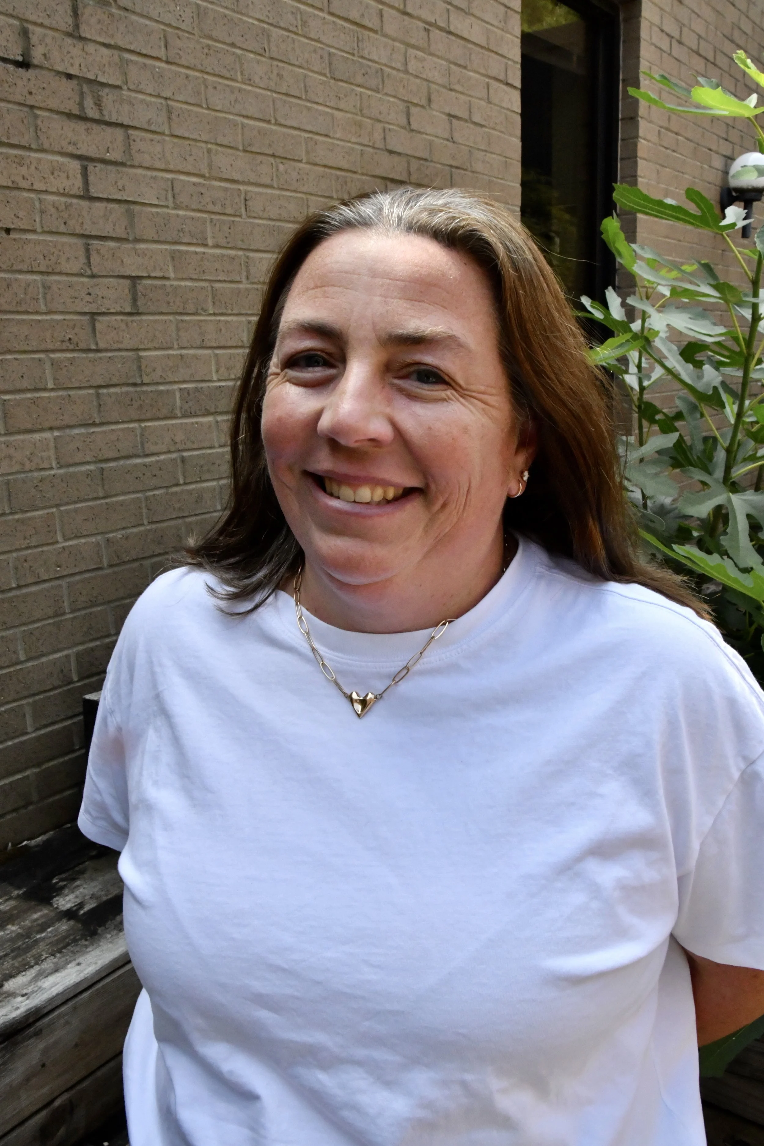 Smiling woman with blonde hair tied back, wearing a white top, standing outdoors with green and yellow foliage in the background.