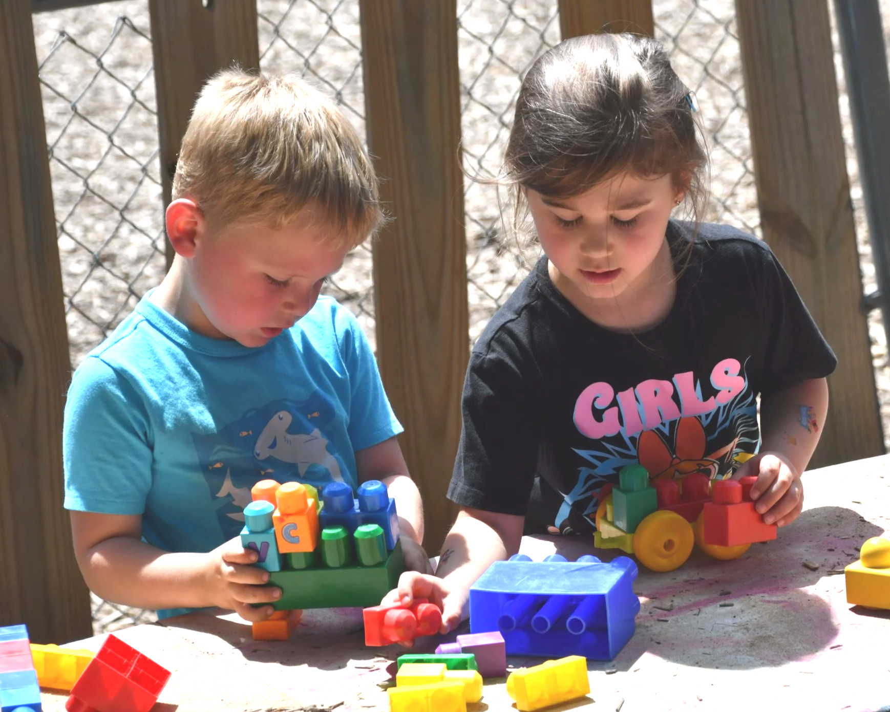 Two children playing with colorful building blocks outdoors on a sunny day, with a wood fence and chain-link fence in the background.
