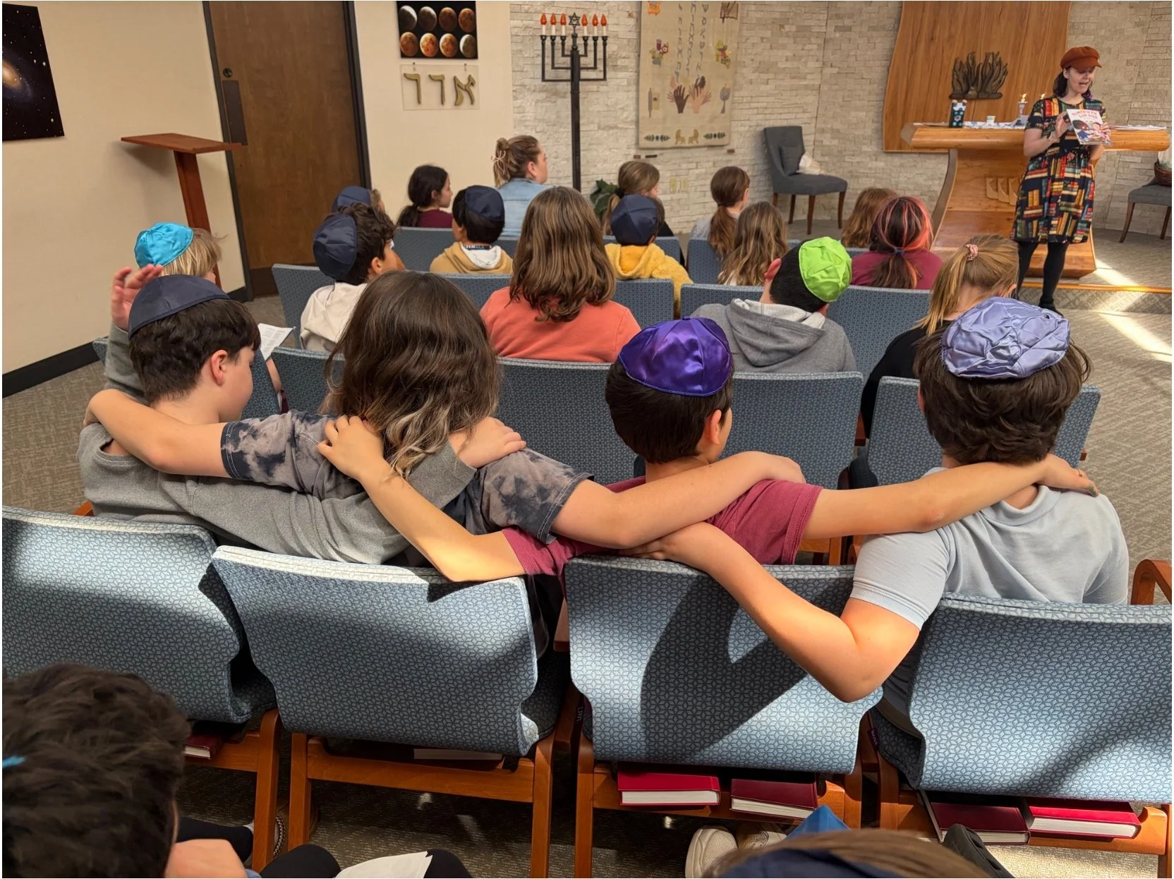 Children sitting in a classroom or meeting room, some wearing kippahs, listening to a woman reading a book at the front near a wooden altar and menorah, with Hebrew symbols and art on the walls.