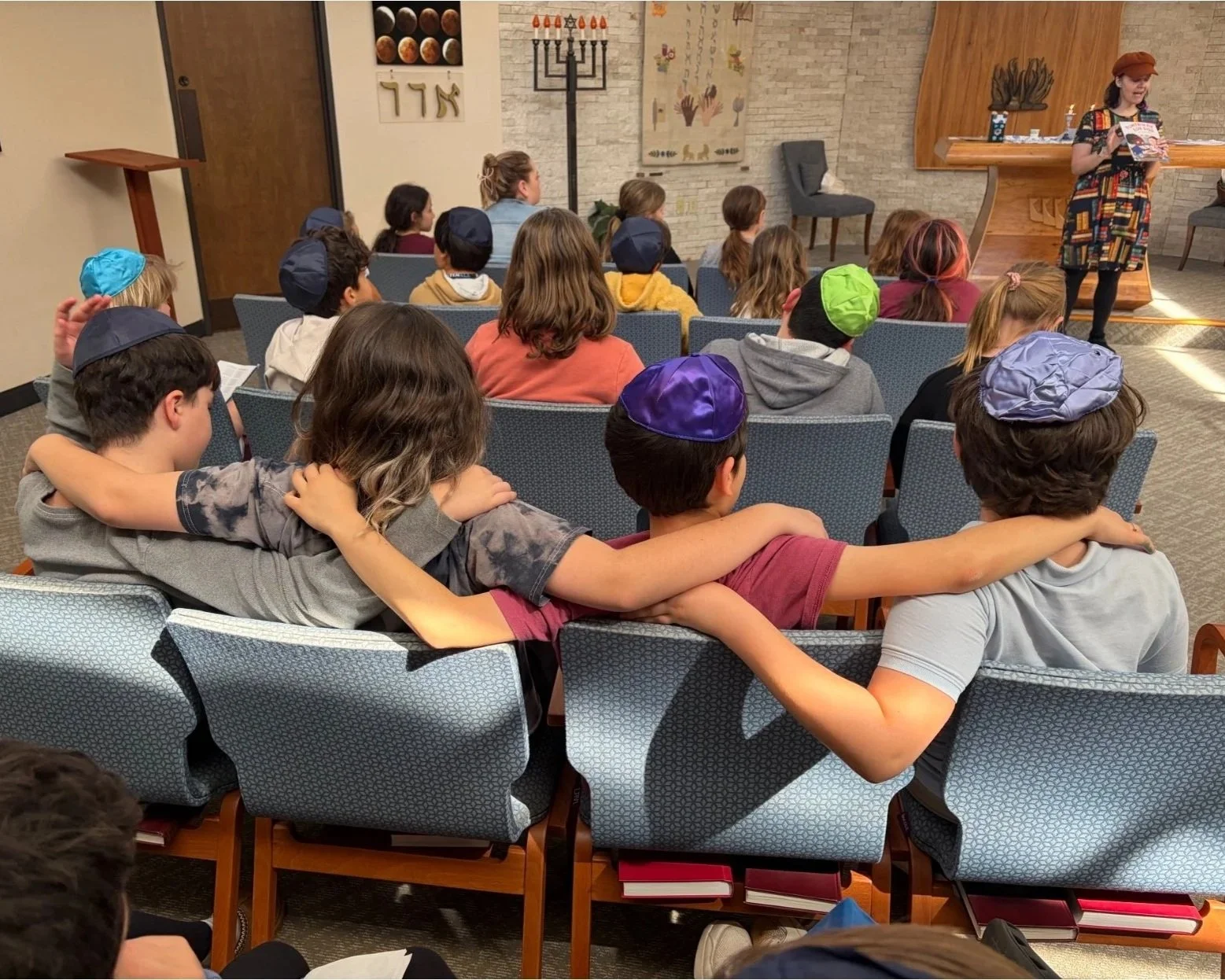A group of children sitting in chairs in a synagogue, with their arms around each other's shoulders, watching a woman reading a book at the front of the room. Some children are wearing kippahs.
