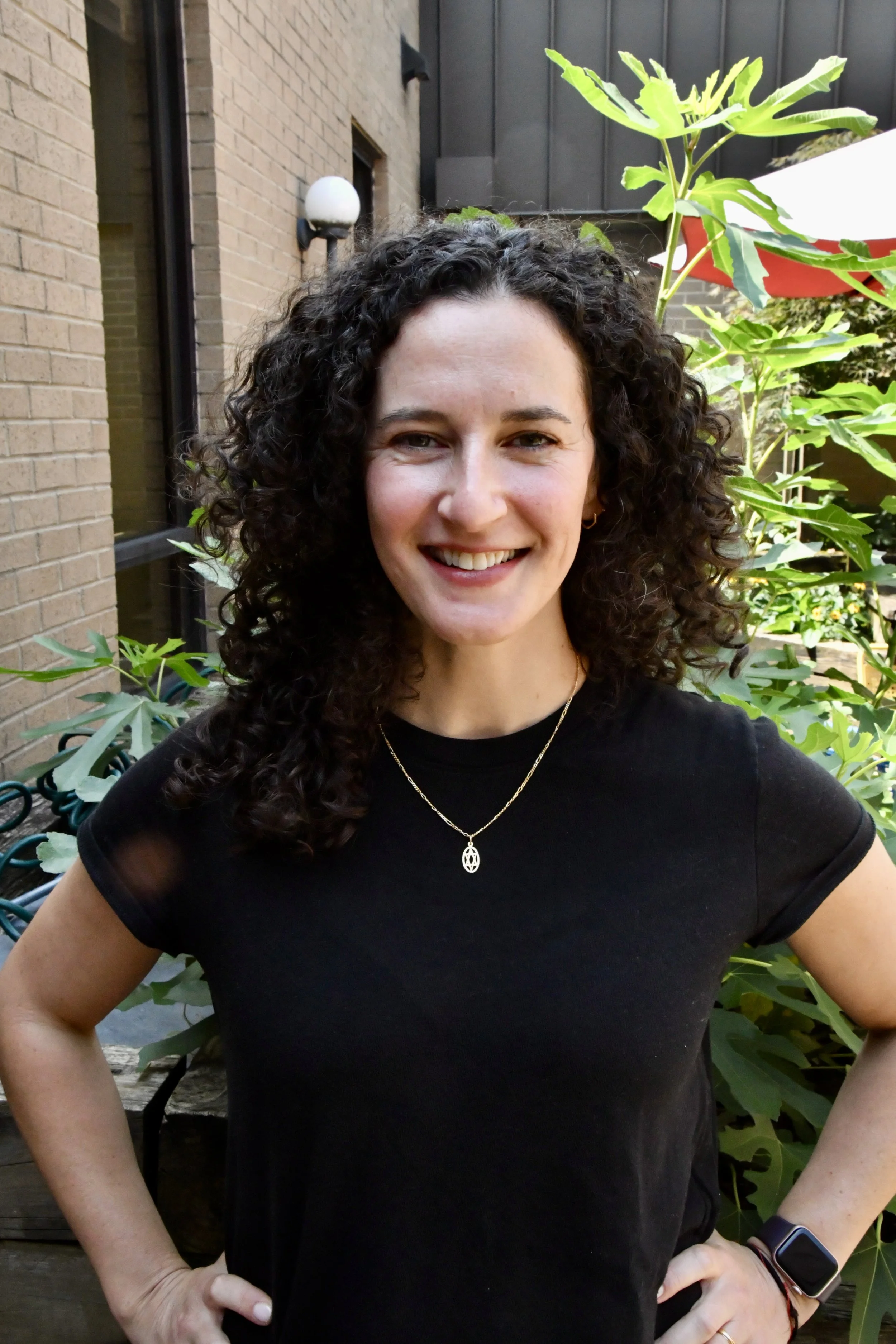 A woman with short curly brown hair smiling outdoors, wearing a green top with floral embroidery, gold necklace, and flower-shaped earrings.