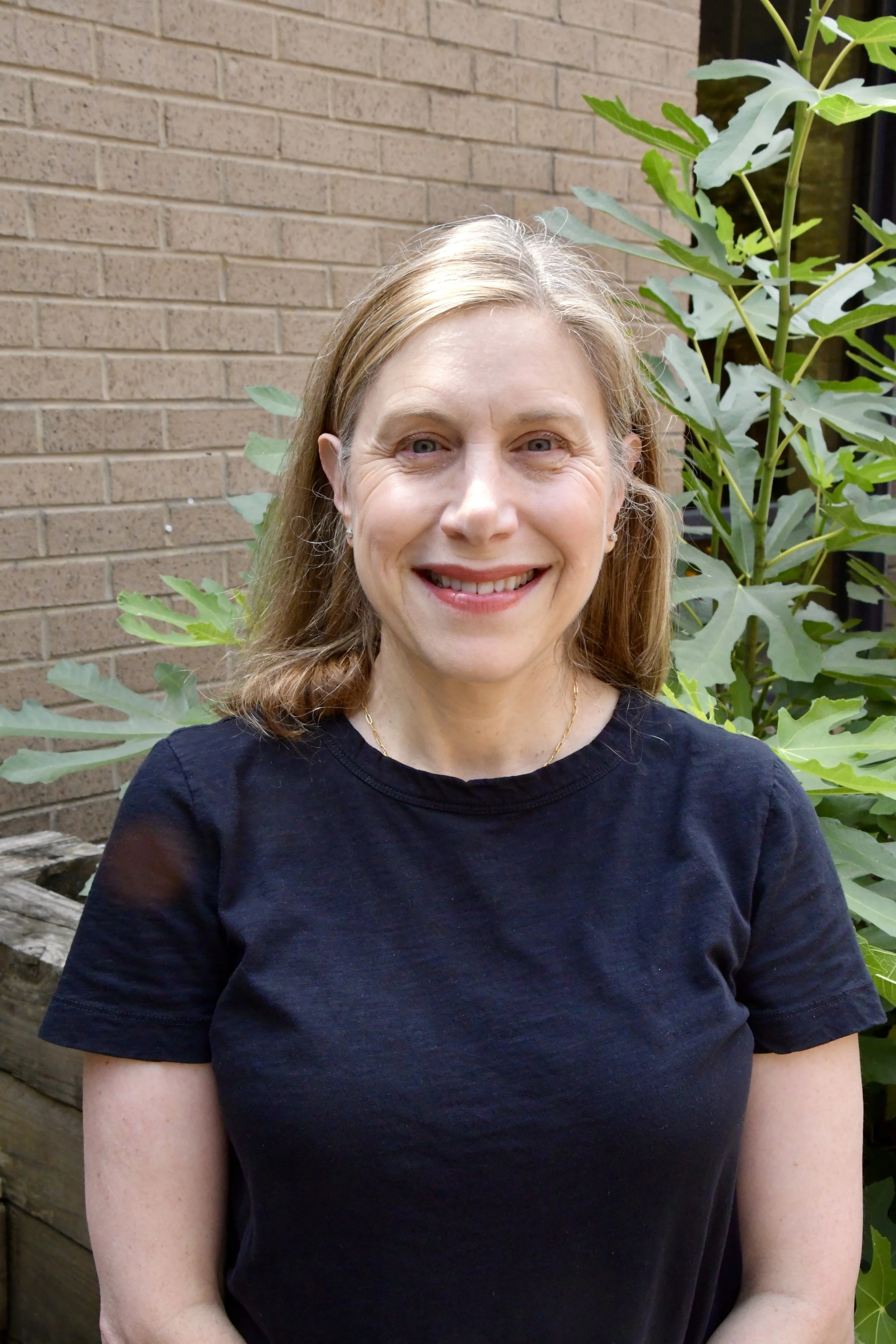 A woman with long, light brown hair and black glasses smiling outdoors with greenery in the background.