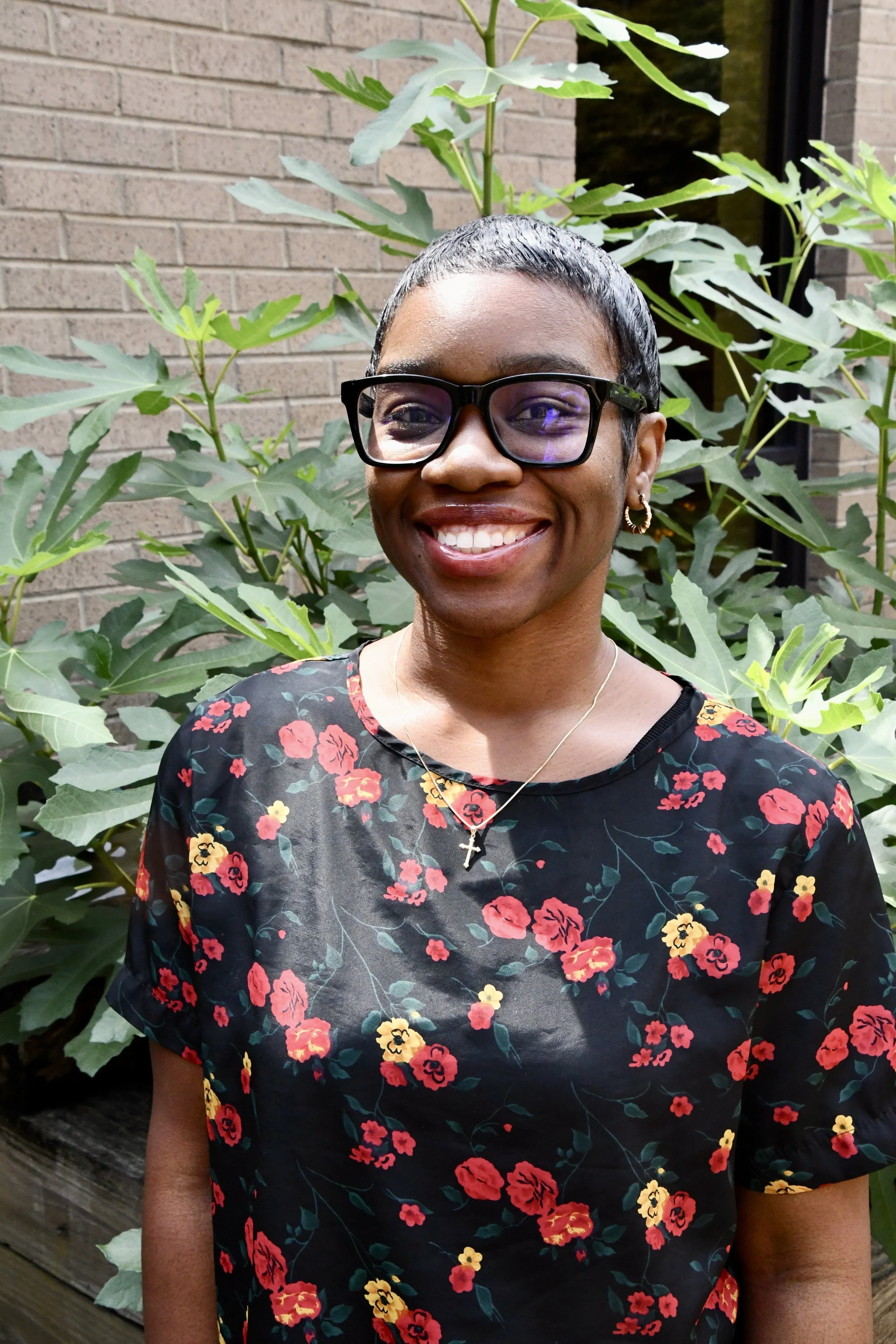 Close-up of a smiling woman with short dark hair wearing round glasses and an orange patterned top, outdoors with blurred trees in the background.
