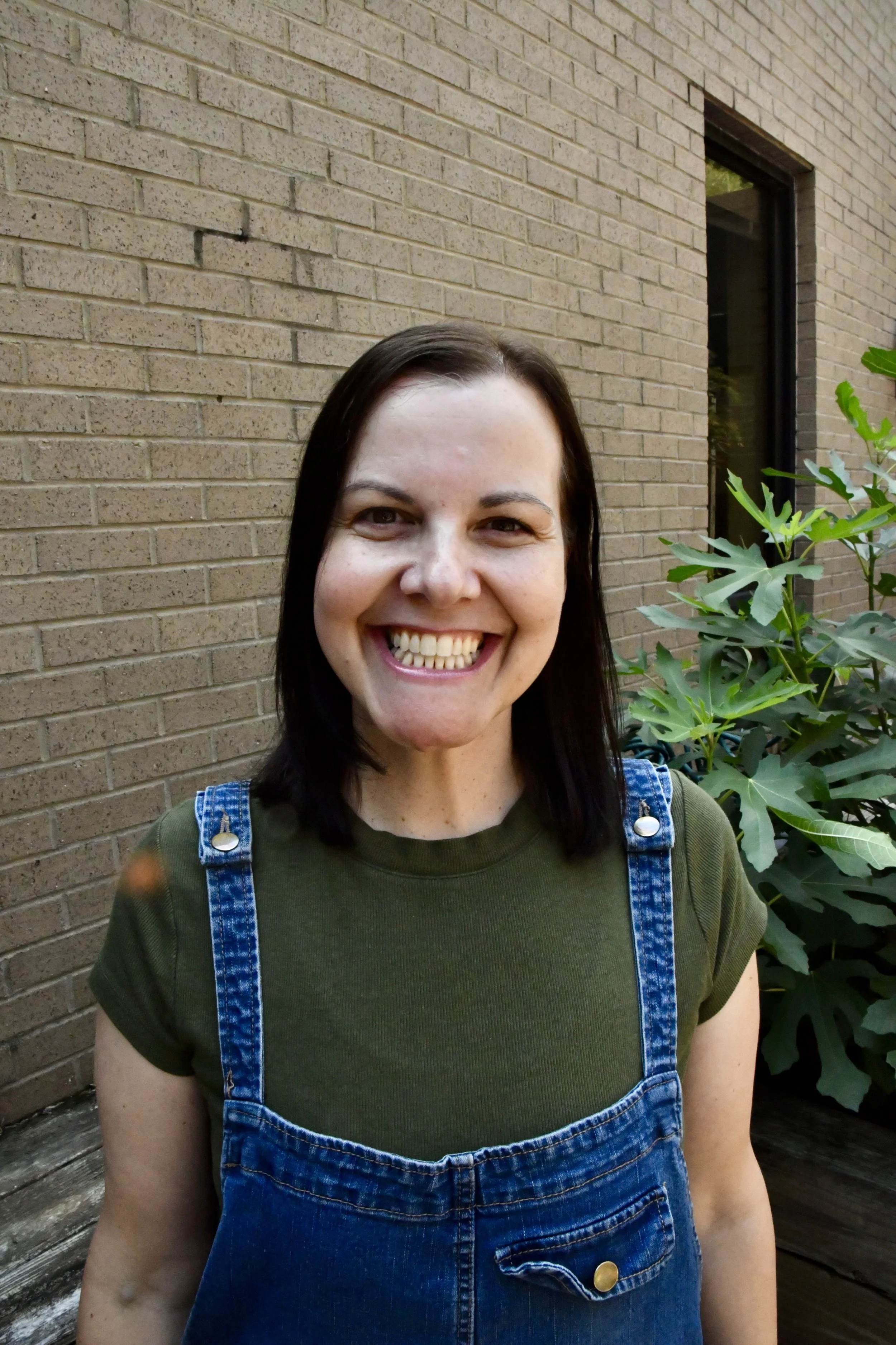 Smiling woman with braces, wearing a red shirt with white trim, standing outdoors with blurry greenery and autumn leaves in the background.
