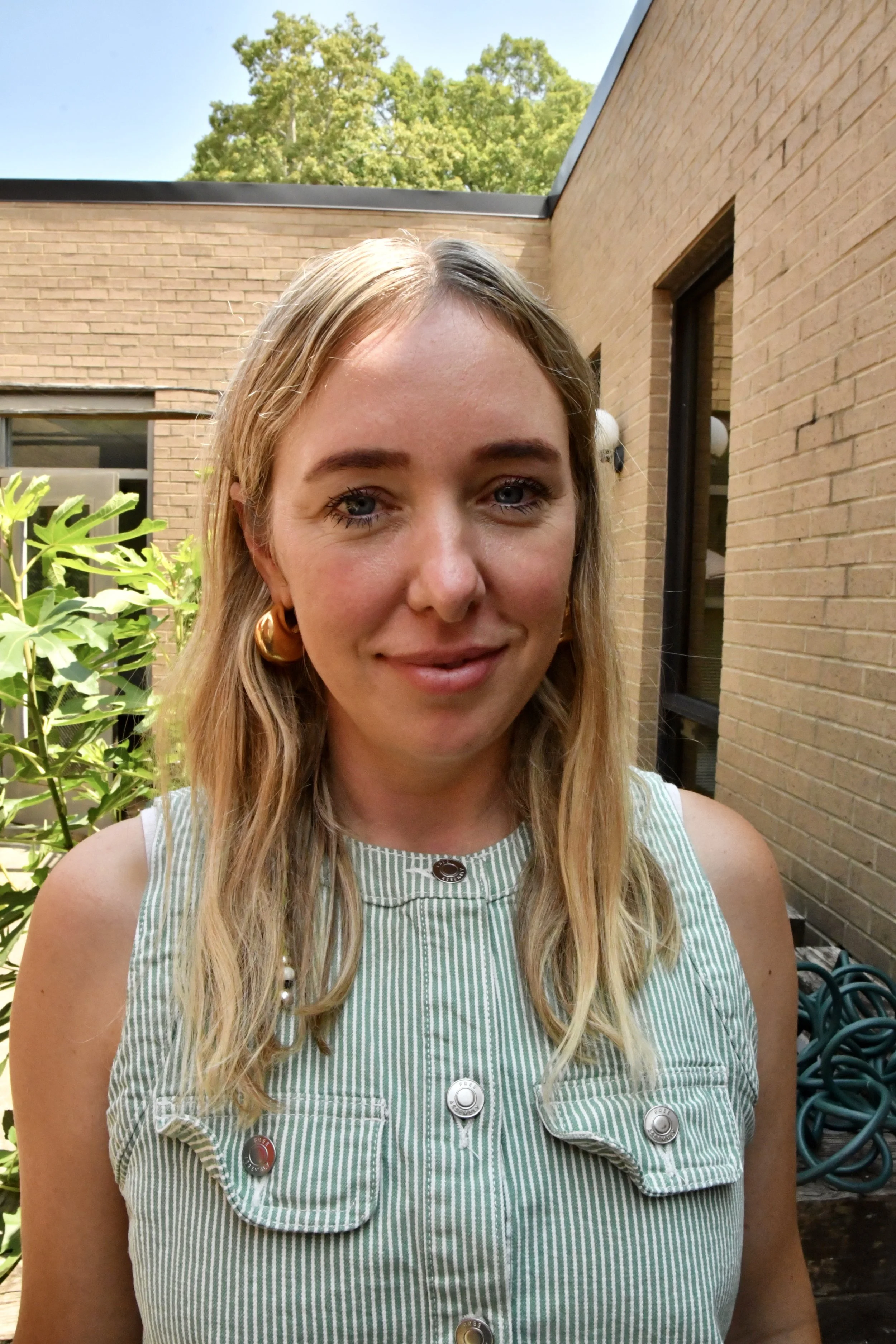 Close-up of a smiling woman with blonde hair, blue eyes, wearing a blue-striped shirt indoors with a blurred natural background.