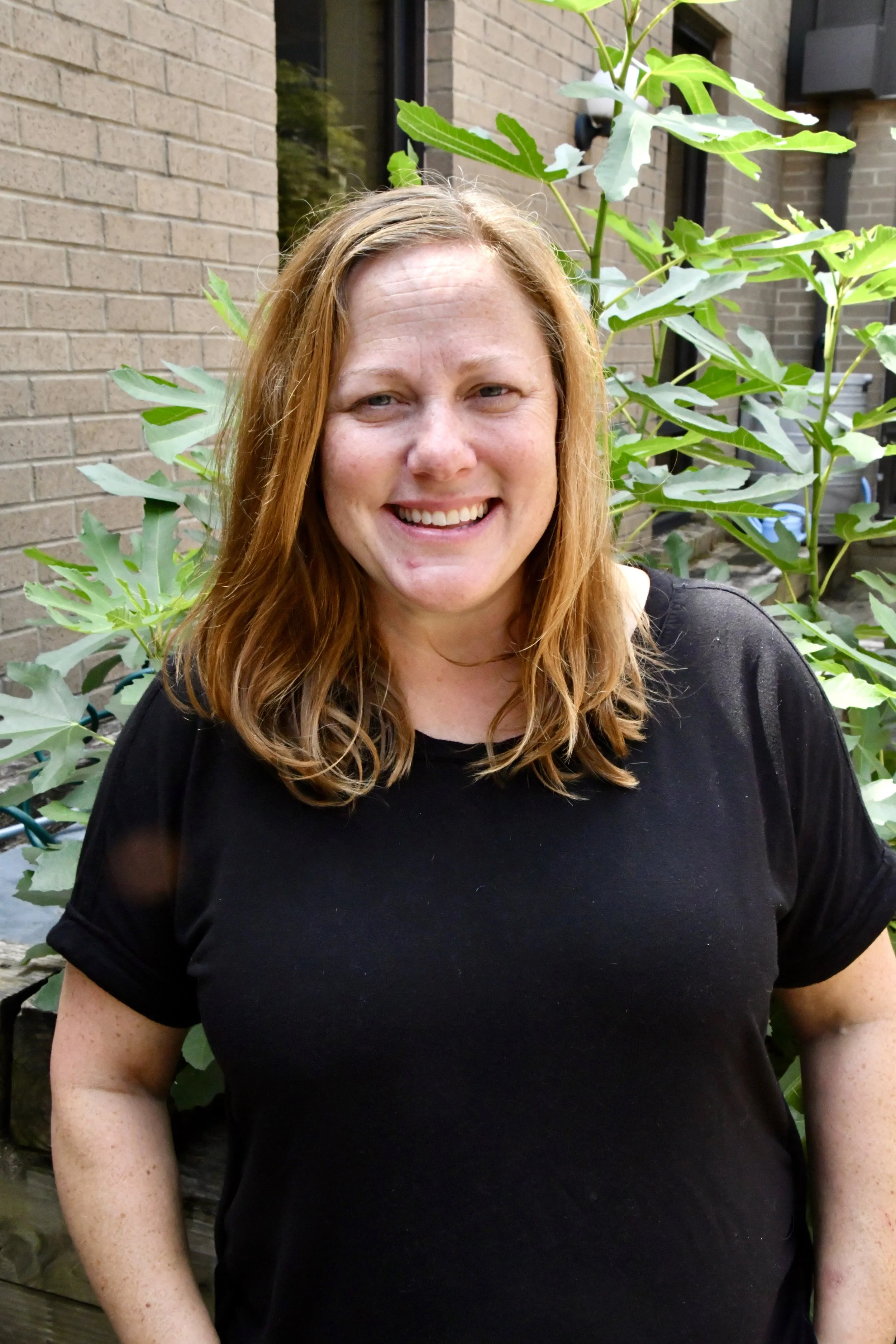 A woman with long red hair smiling outdoors near trees.