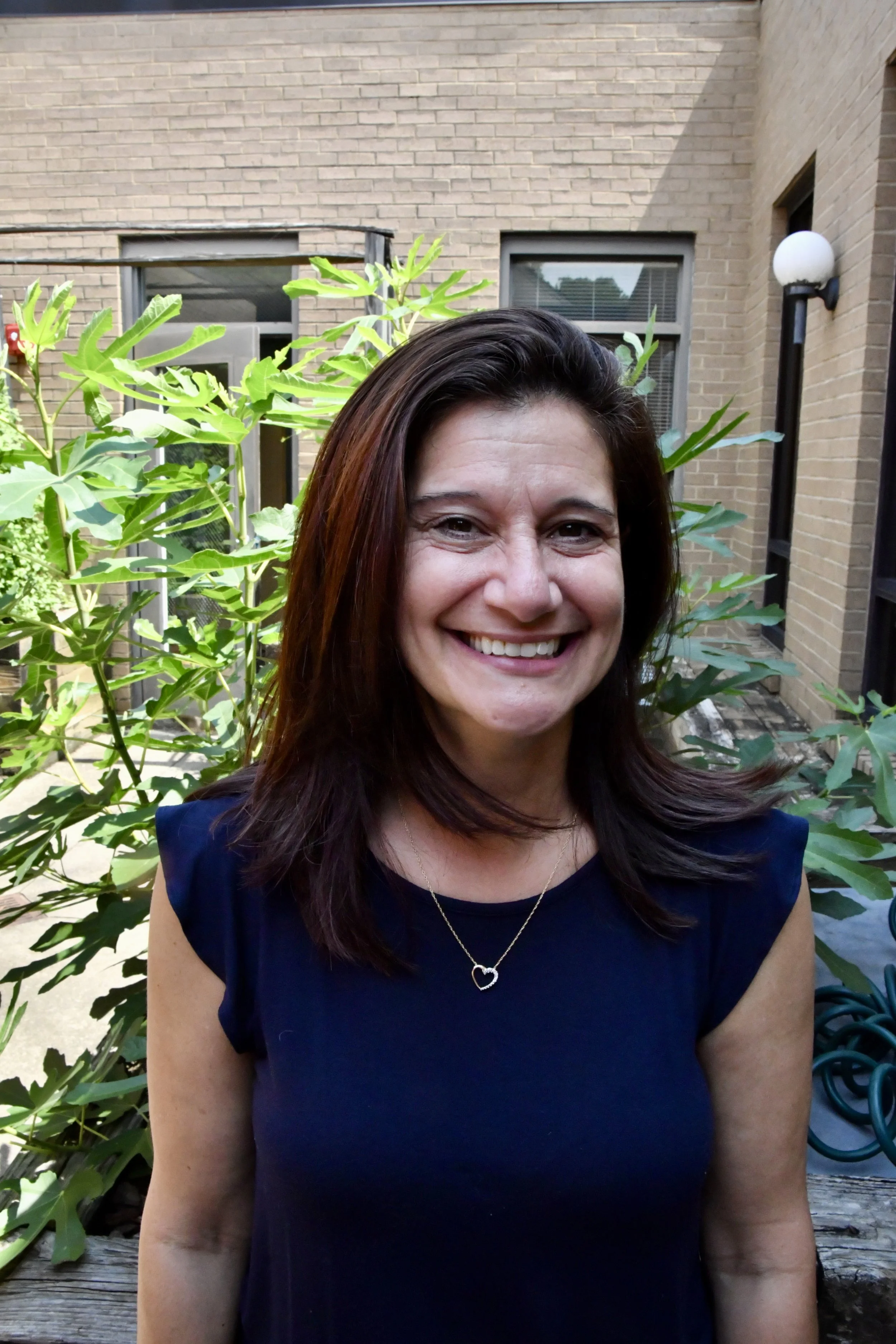A smiling woman with shoulder-length brown hair, wearing a blue top and a necklace, standing outdoors with greenery and foliage in the background.