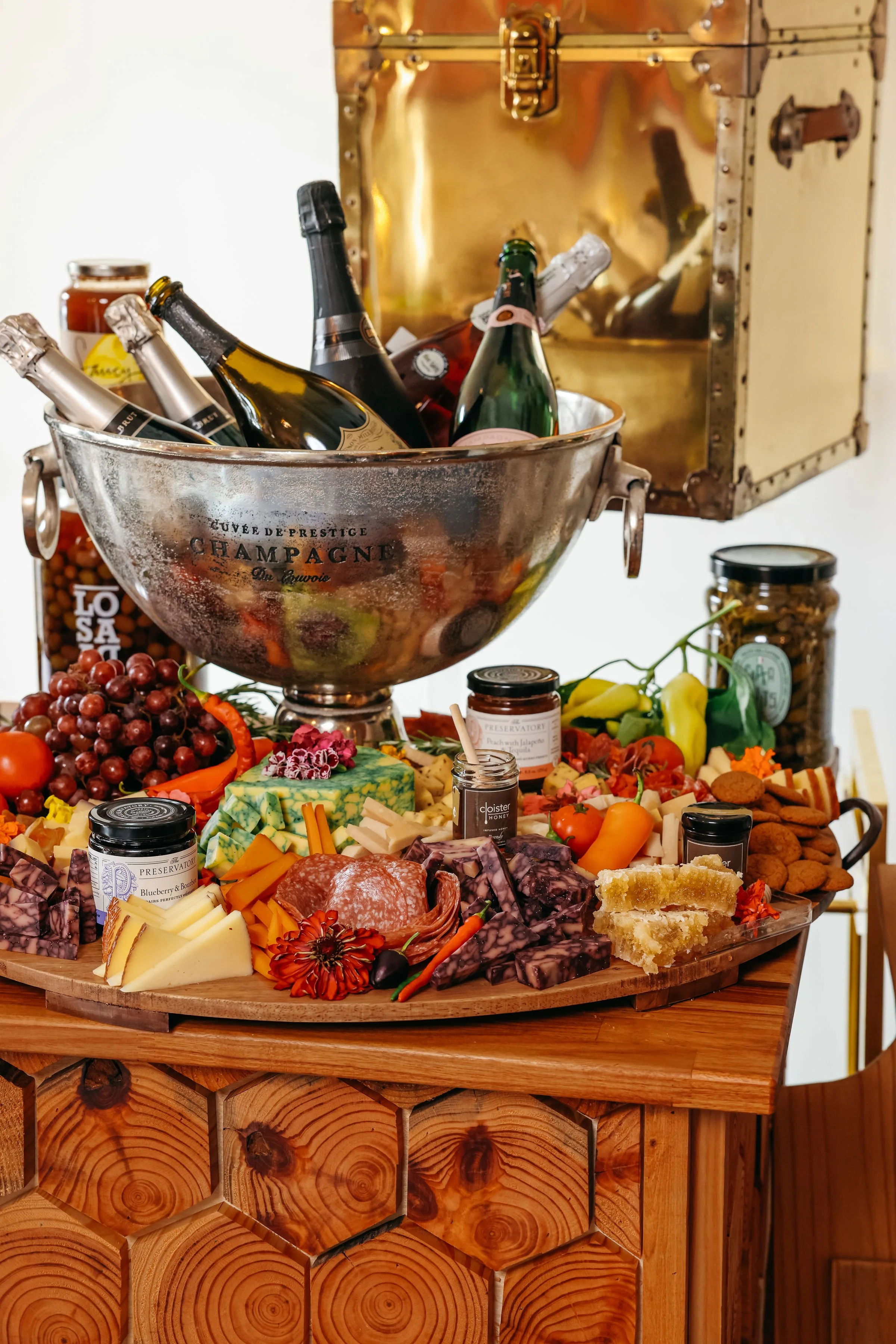 A cheese and charcuterie board with grapes, vegetables, and chocolate, topped with bottles of wine and champagne in an ice bucket, on a wooden table with jars of preserves in the background.