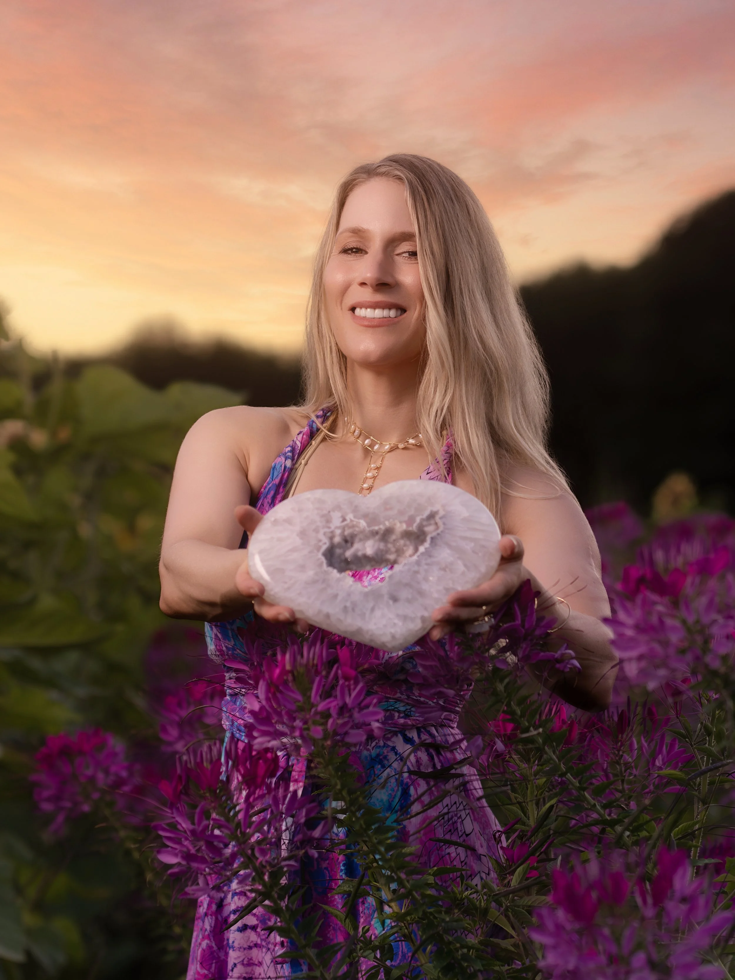 Carine A-Hamid offrant un cœur d’améthyste rose, entourée de fleurs pourpres sous un ciel aux nuances pêche et rosées.