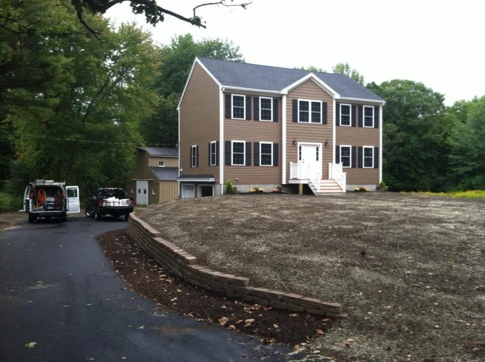 Newly constructed three-story house with brown siding, black shutters, and a white front door, surrounded by a yard with dirt and a brick border, and two vehicles parked on a driveway to the left, with trees in the background.