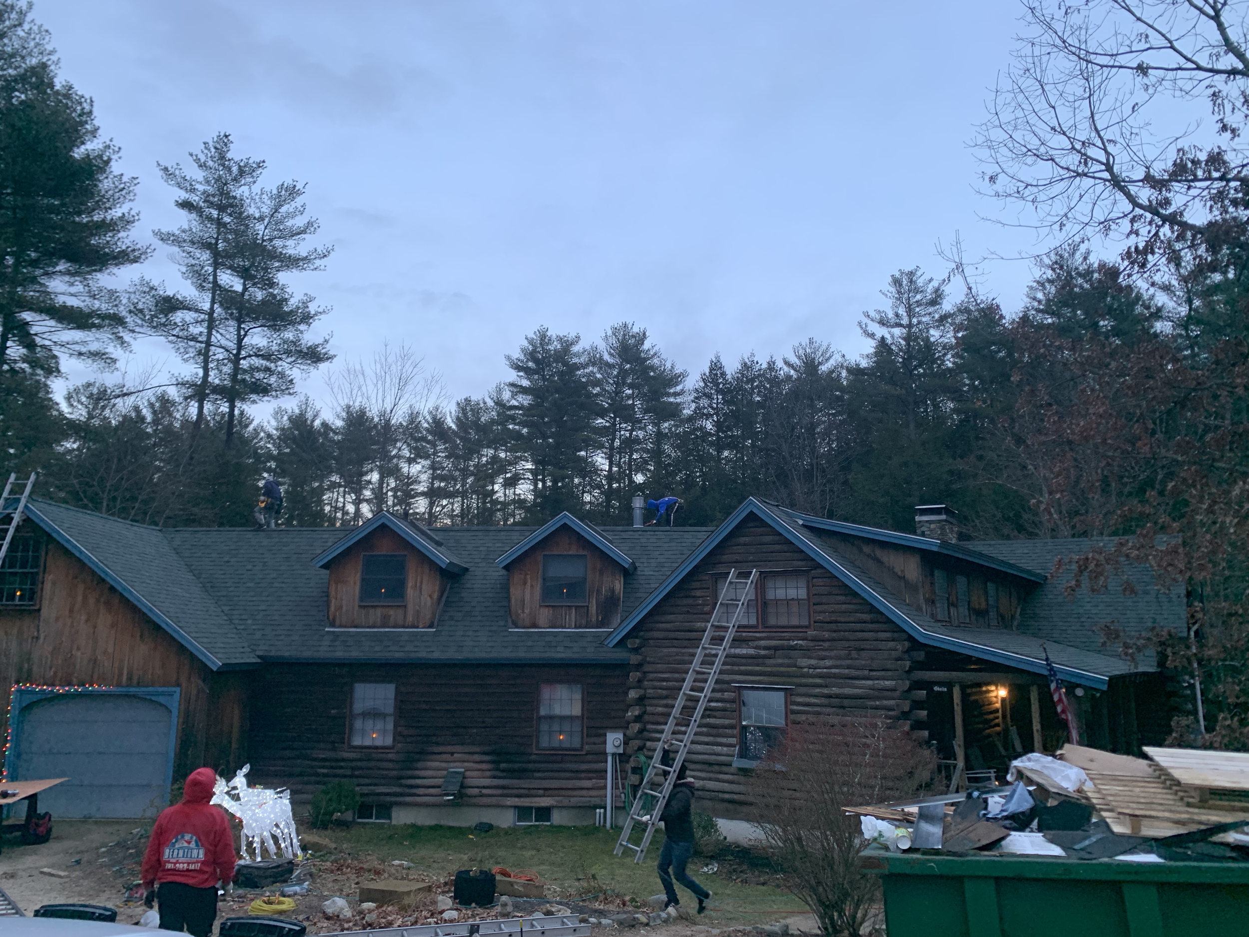 Workers on the roof of a house with a dark wood exterior, surrounded by trees, with equipment and workers below.