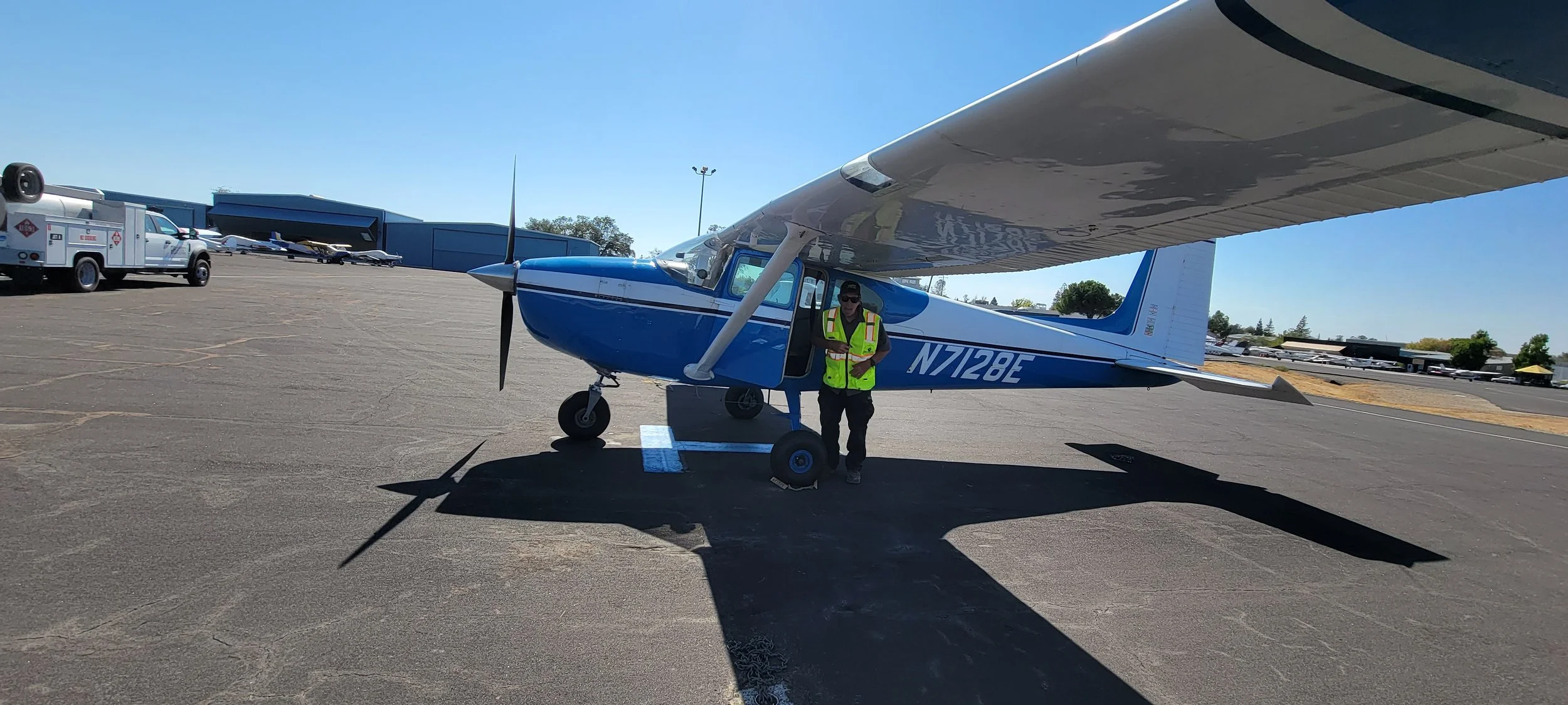 Blue single-engine airplane on a tarmac with a person in a safety vest and a white fuel truck nearby, under a clear blue sky.