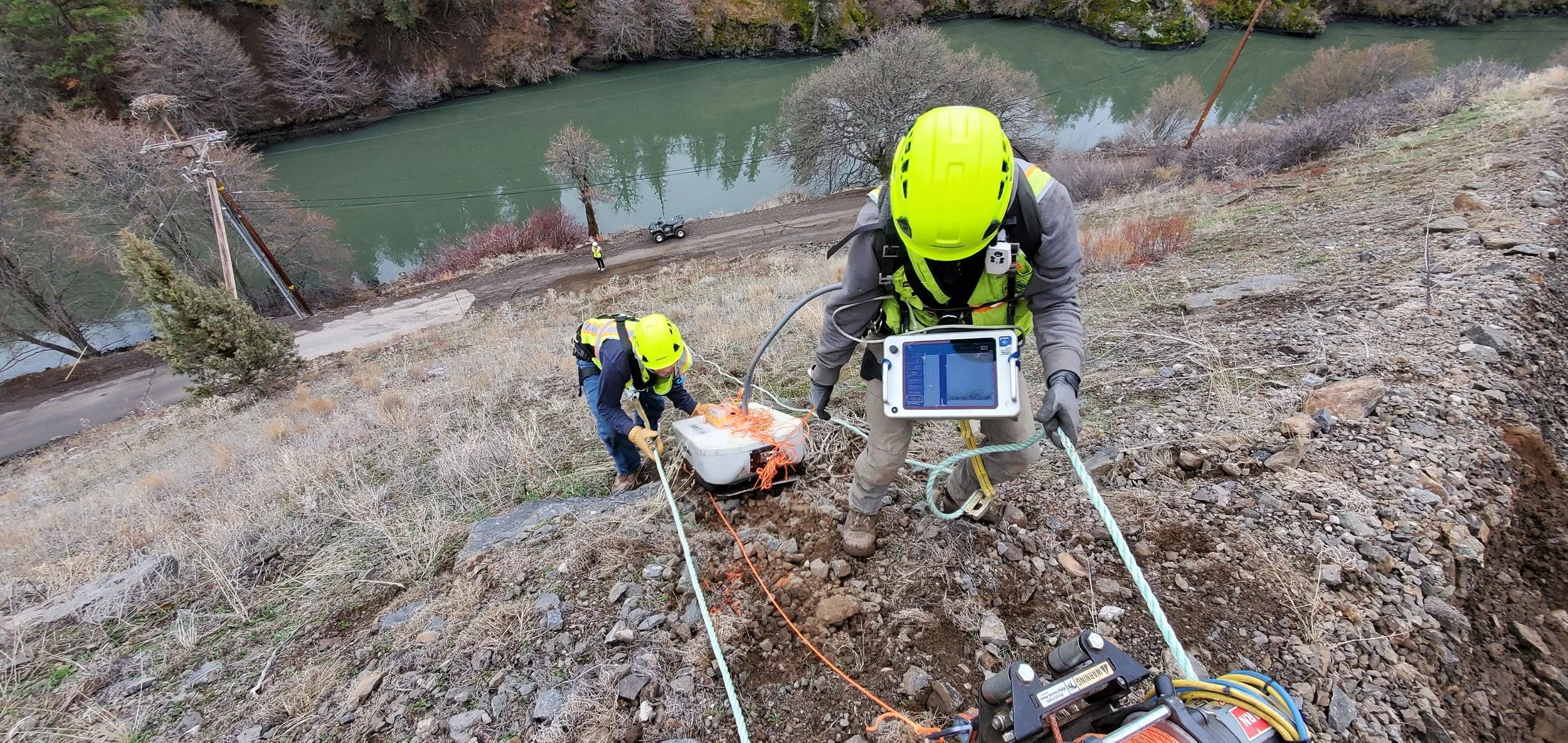 Two workers in helmets conducting a geotechnical survey on a steep slope near a river.