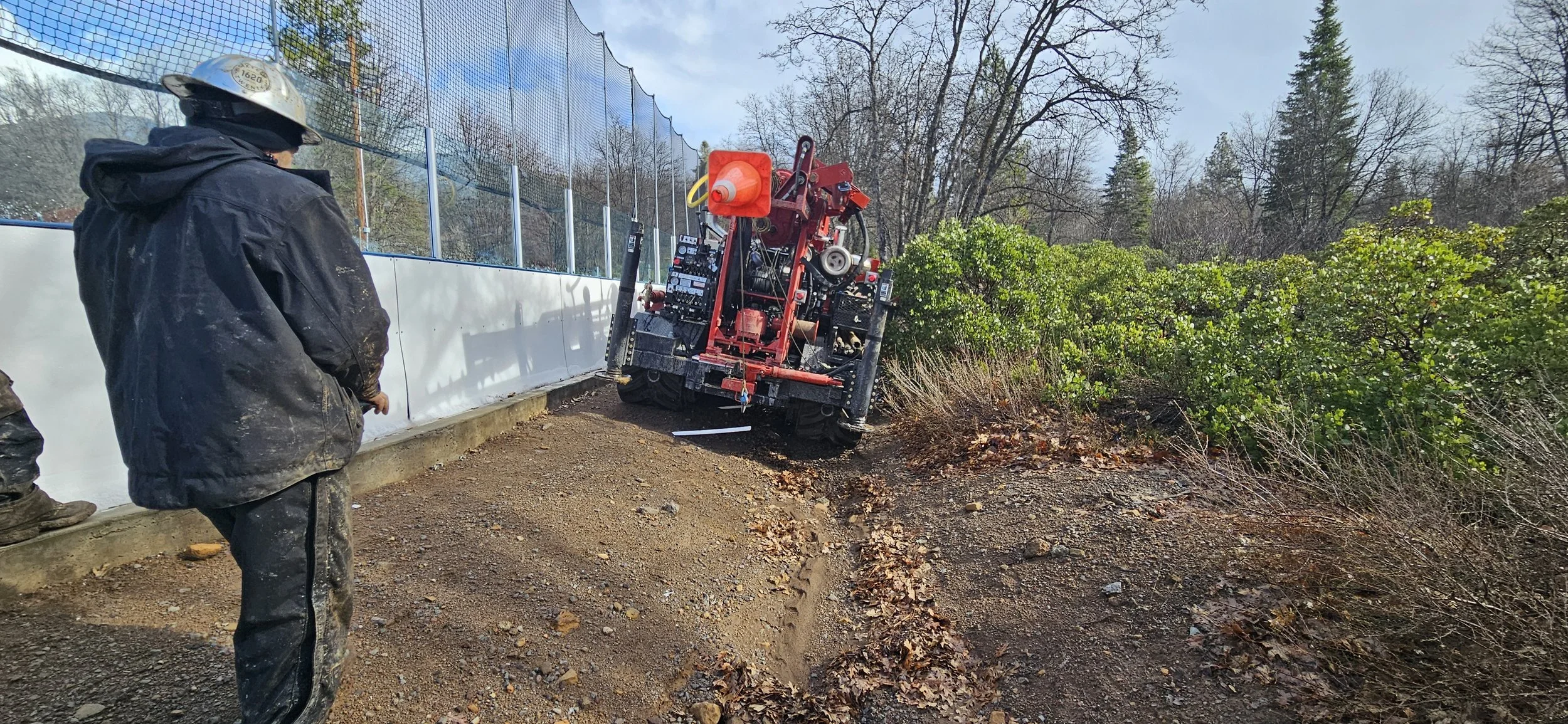 Worker in black jacket and helmet observing a red machine stuck on the edge of a dirt path near a mesh fence, surrounded by trees and shrubs.