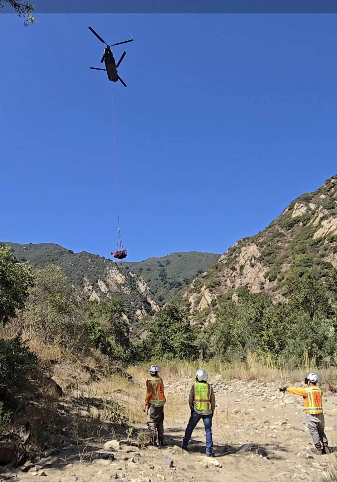 helicopter lifting cargo in a mountainous area with people observing