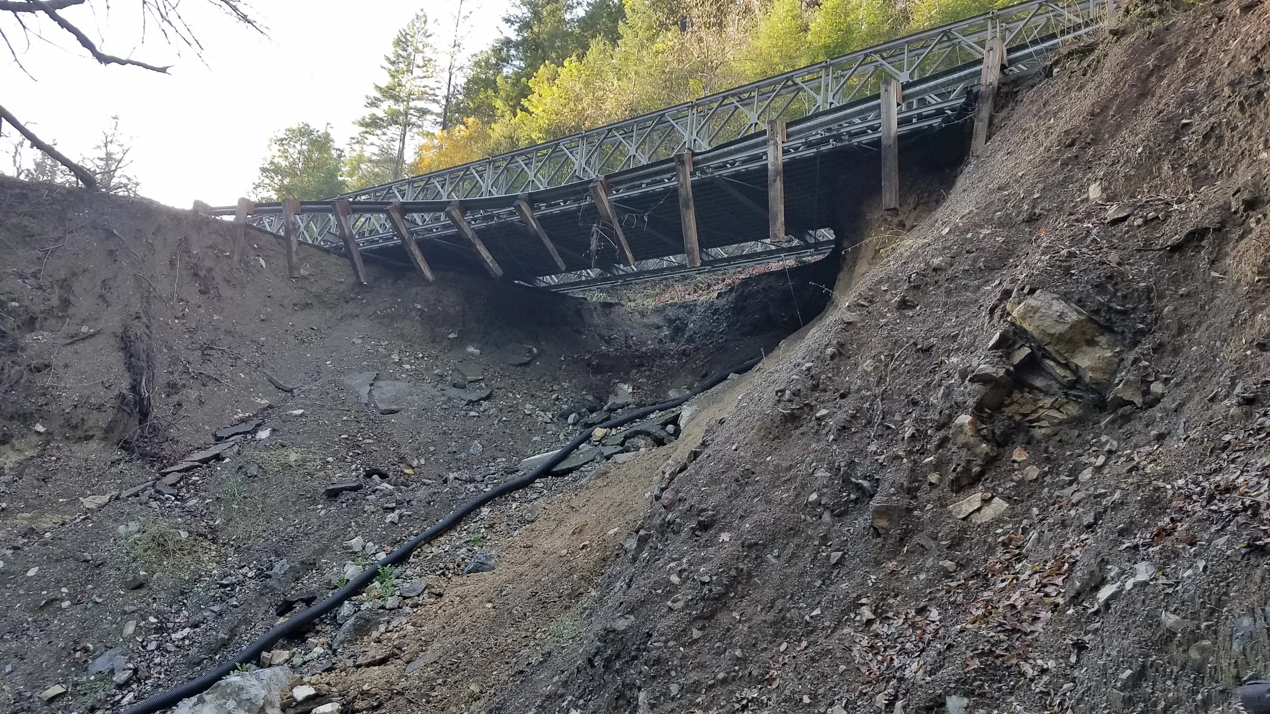 Old damaged bridge with visible erosion and exposed dirt beneath, alongside forested area.