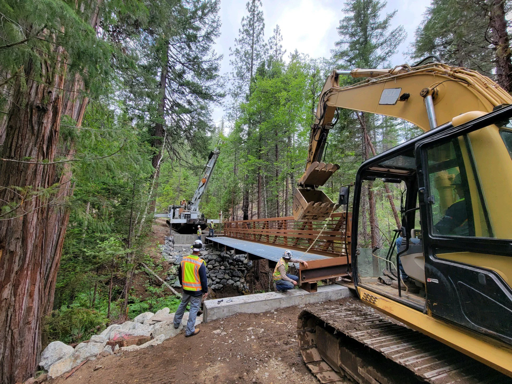 Construction workers using heavy machinery to build a bridge in a forested area.