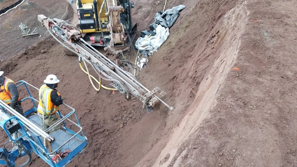 Construction workers in orange vests and white hard hats operating machinery on a dirt slope.