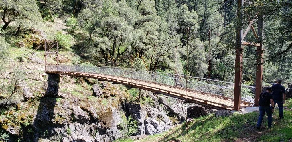 Suspension bridge over a rocky gorge in a forested area, with two people walking towards it.