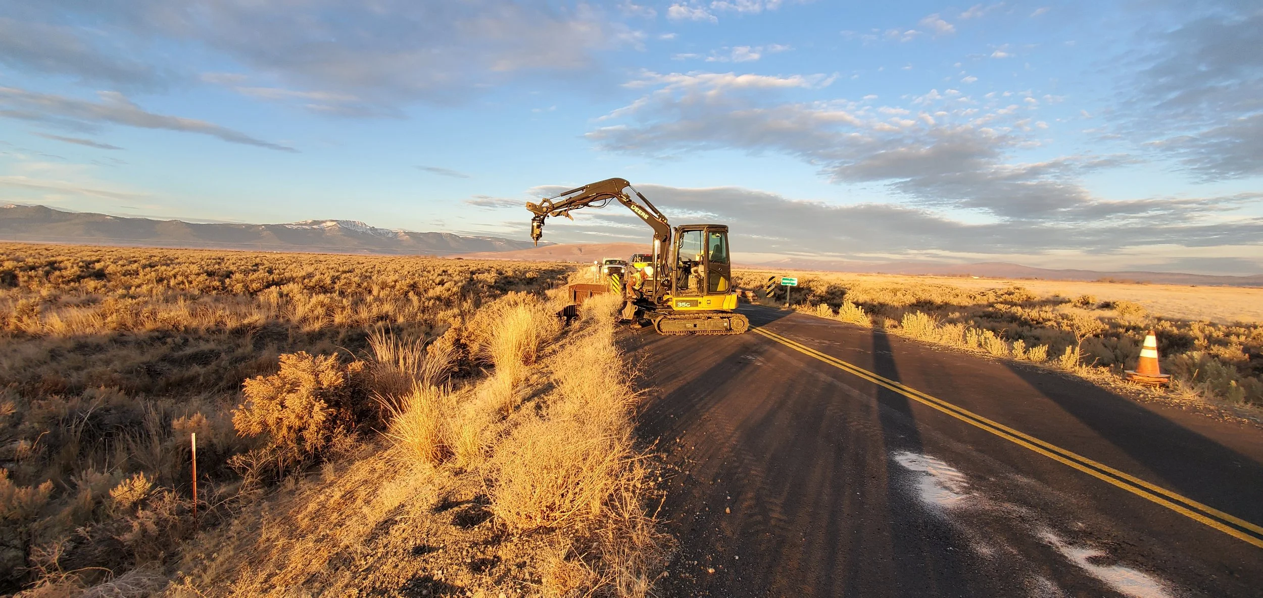 Excavator on rural road with desert landscape and mountains, construction cones, and sky with clouds.