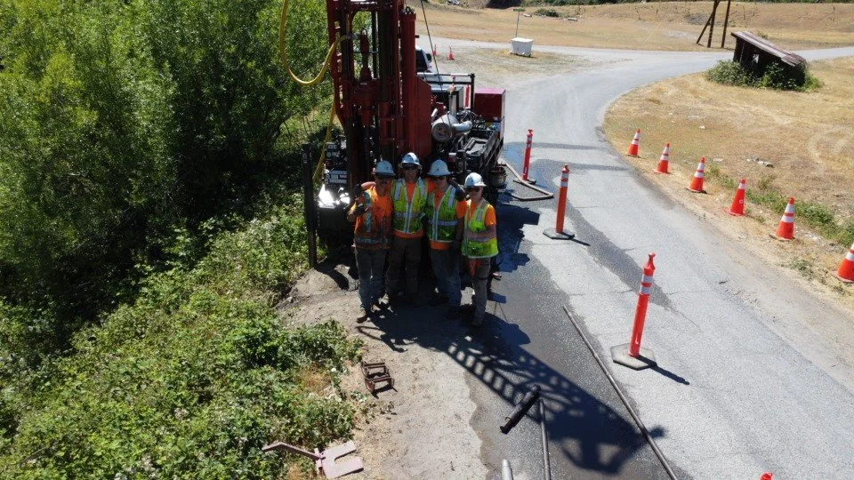 Construction workers in safety gear standing near drilling equipment on a road, surrounded by traffic cones and greenery.