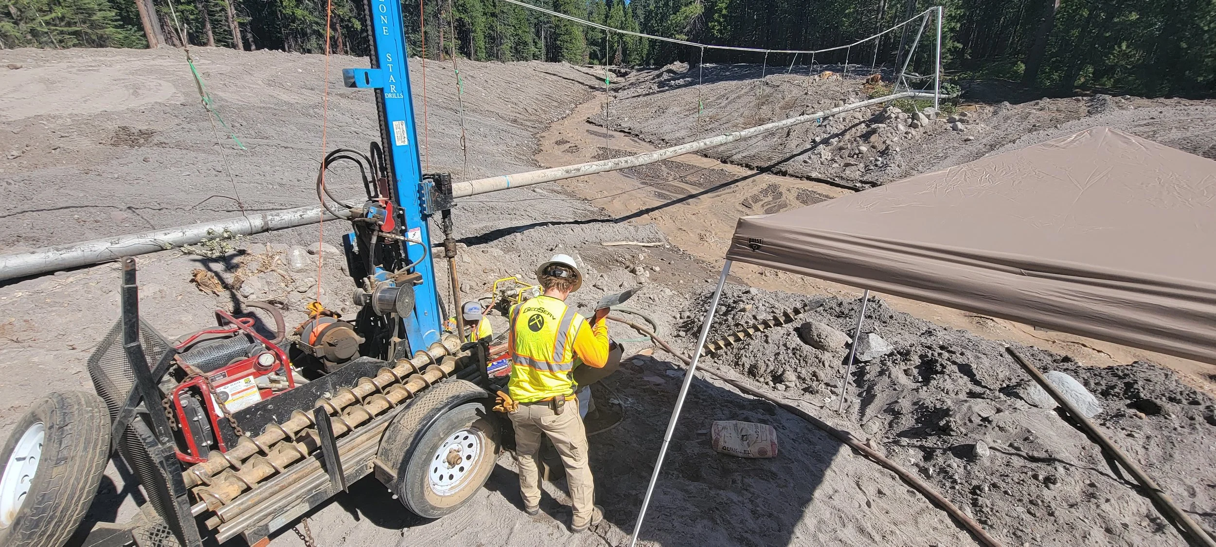 Construction worker at a drilling site with equipment and pipes in an outdoor, forested area.