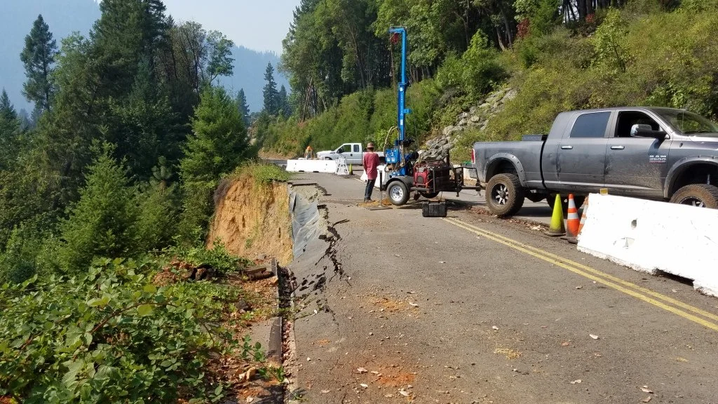Landslide on forest road with damaged pavement, construction vehicles, and barriers.