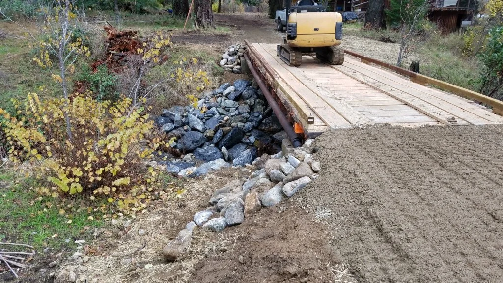 Small bridge construction over rocks with a bulldozer on top, surrounded by trees and bushes.